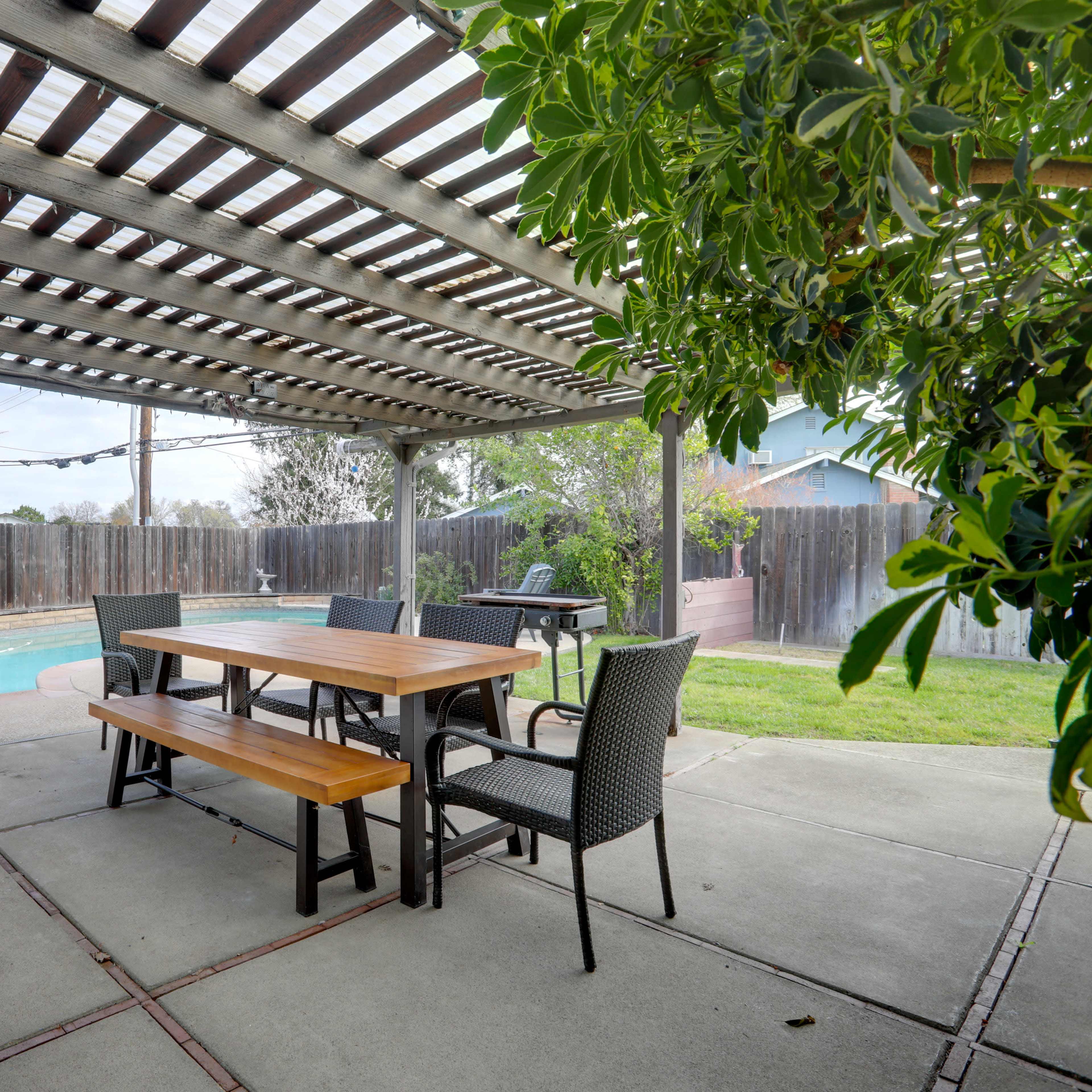 A patio area with a wooden table and benches beneath a pergola, adjacent to a swimming pool and surrounded by a grassy yard.