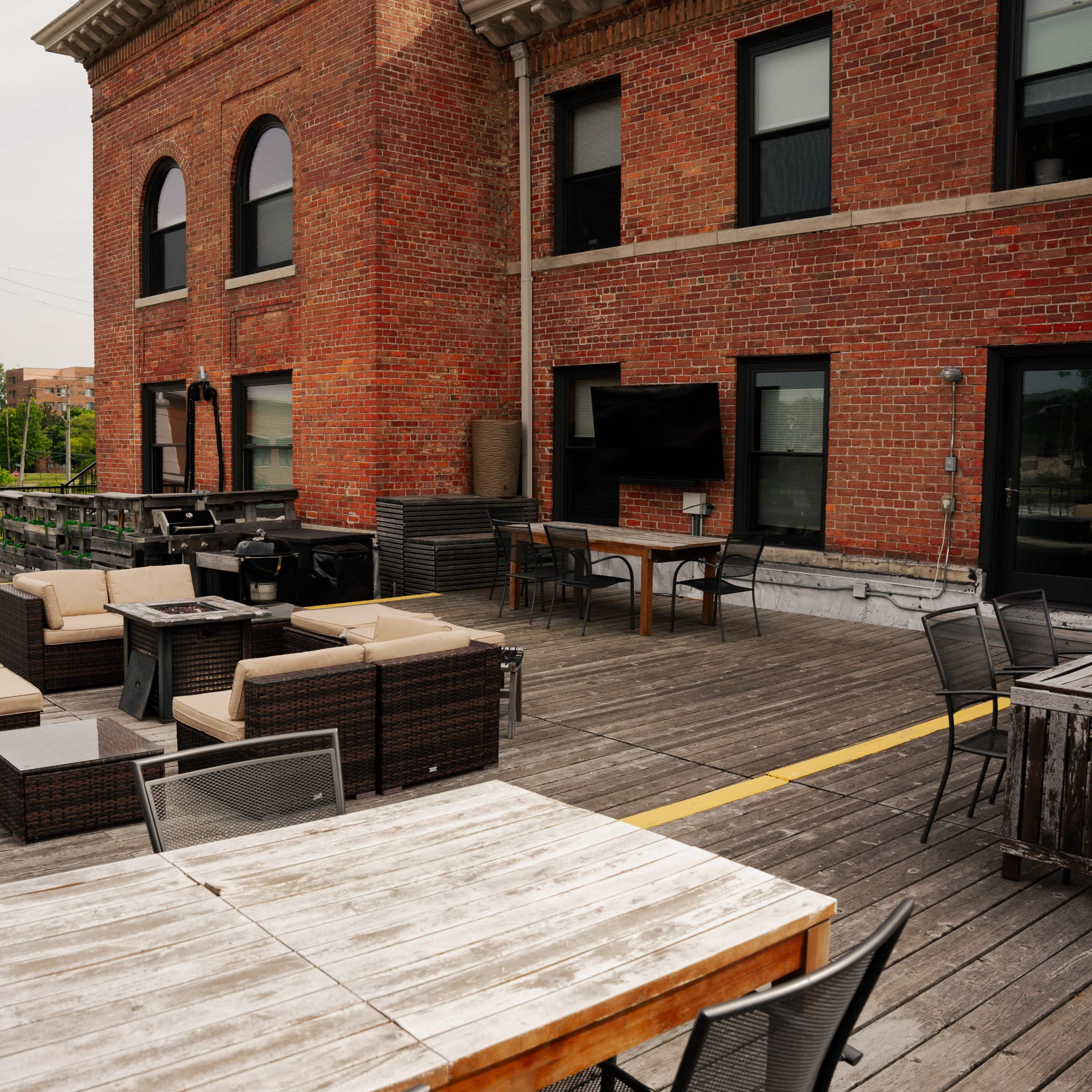 A spacious outdoor patio area with wicker furniture, wooden tables, and a large brick building in the background.