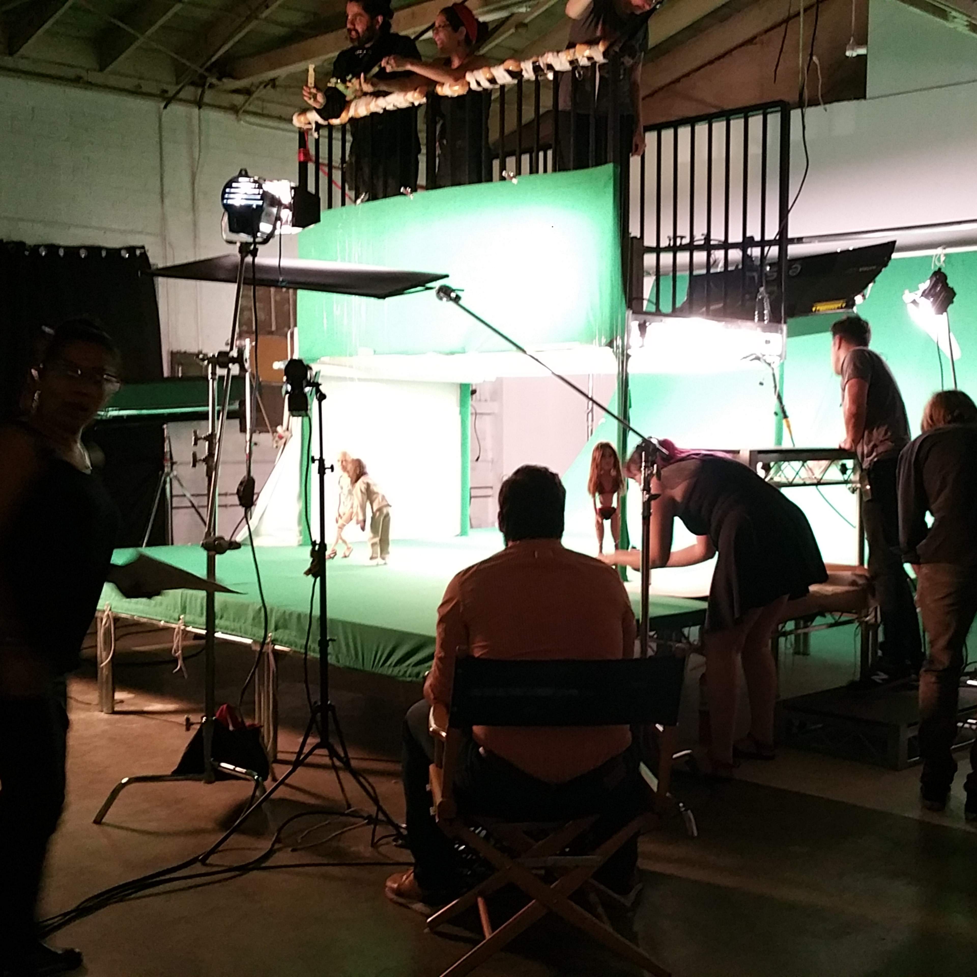 A film crew works in a studio setting with a green screen backdrop, adjusting lights and equipment.
