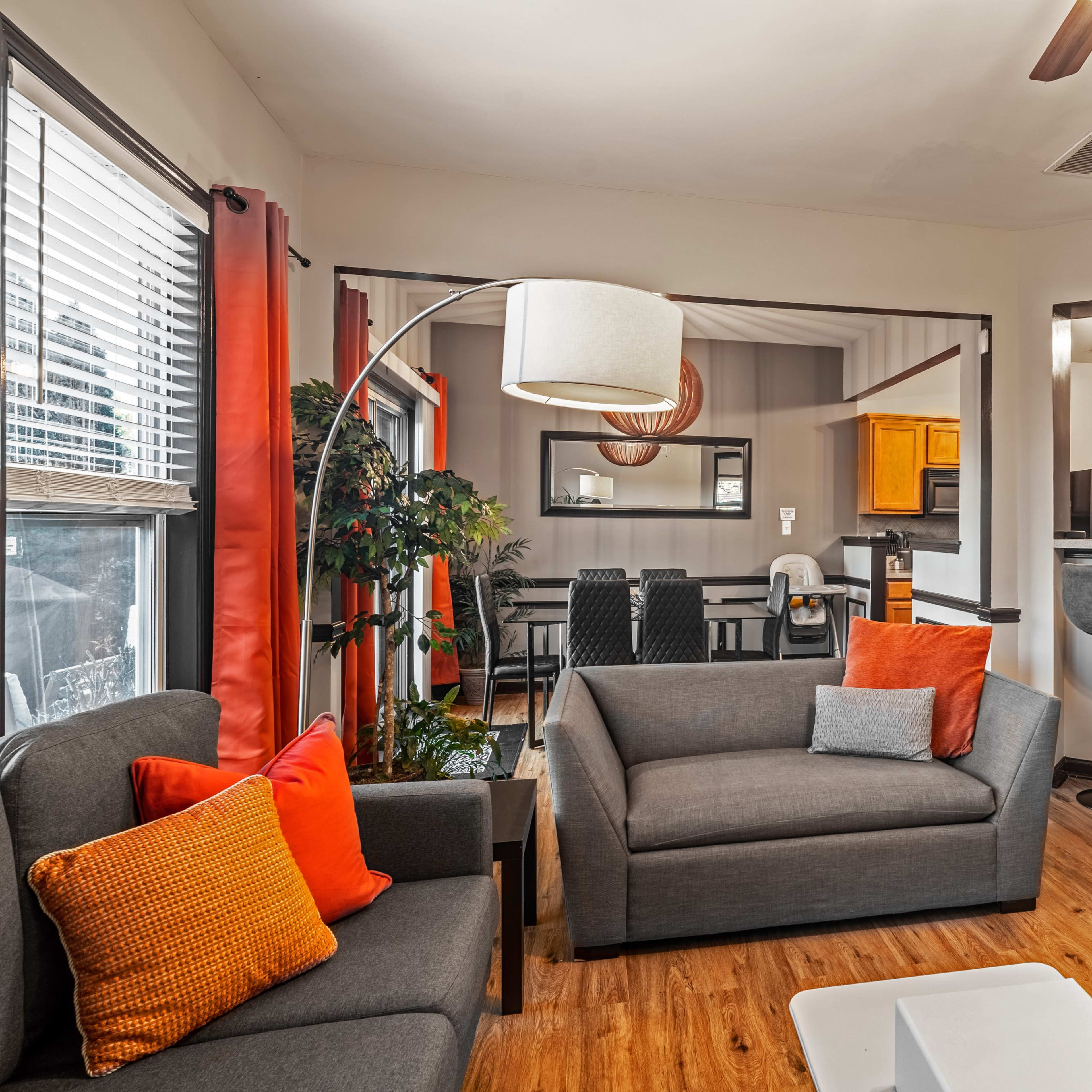 A living room area with a gray sofa, an orange accent chair, a floor lamp, and a view into a dining area with a kitchen in the background.