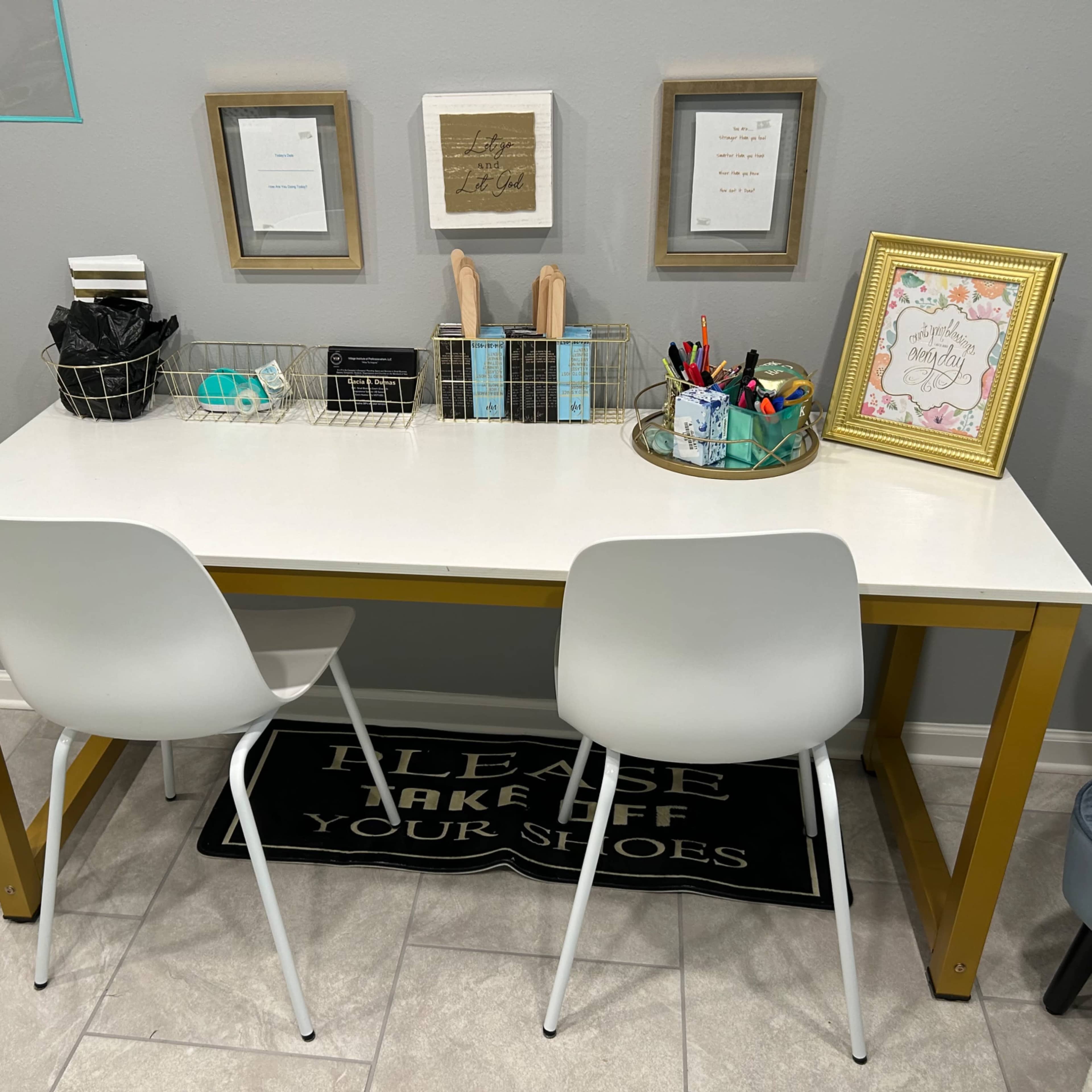 A modern desk with gold legs is adorned with various stationery items and framed pictures, accompanied by two white chairs and a "Please Take Off Your Shoes" rug.