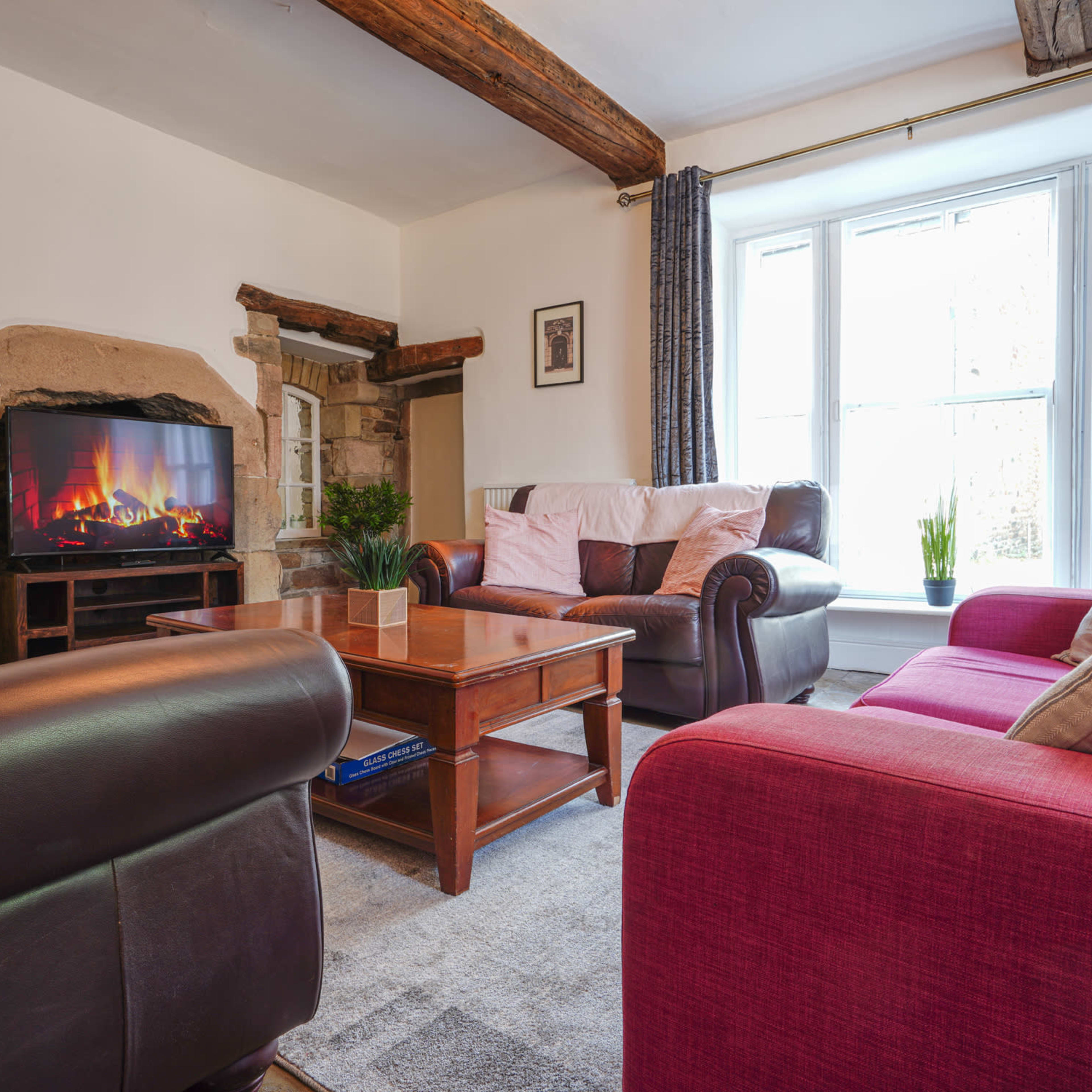 A cozy living room with leather sofas, a wooden coffee table, and a television displaying a fireplace, situated by a window with natural light.
