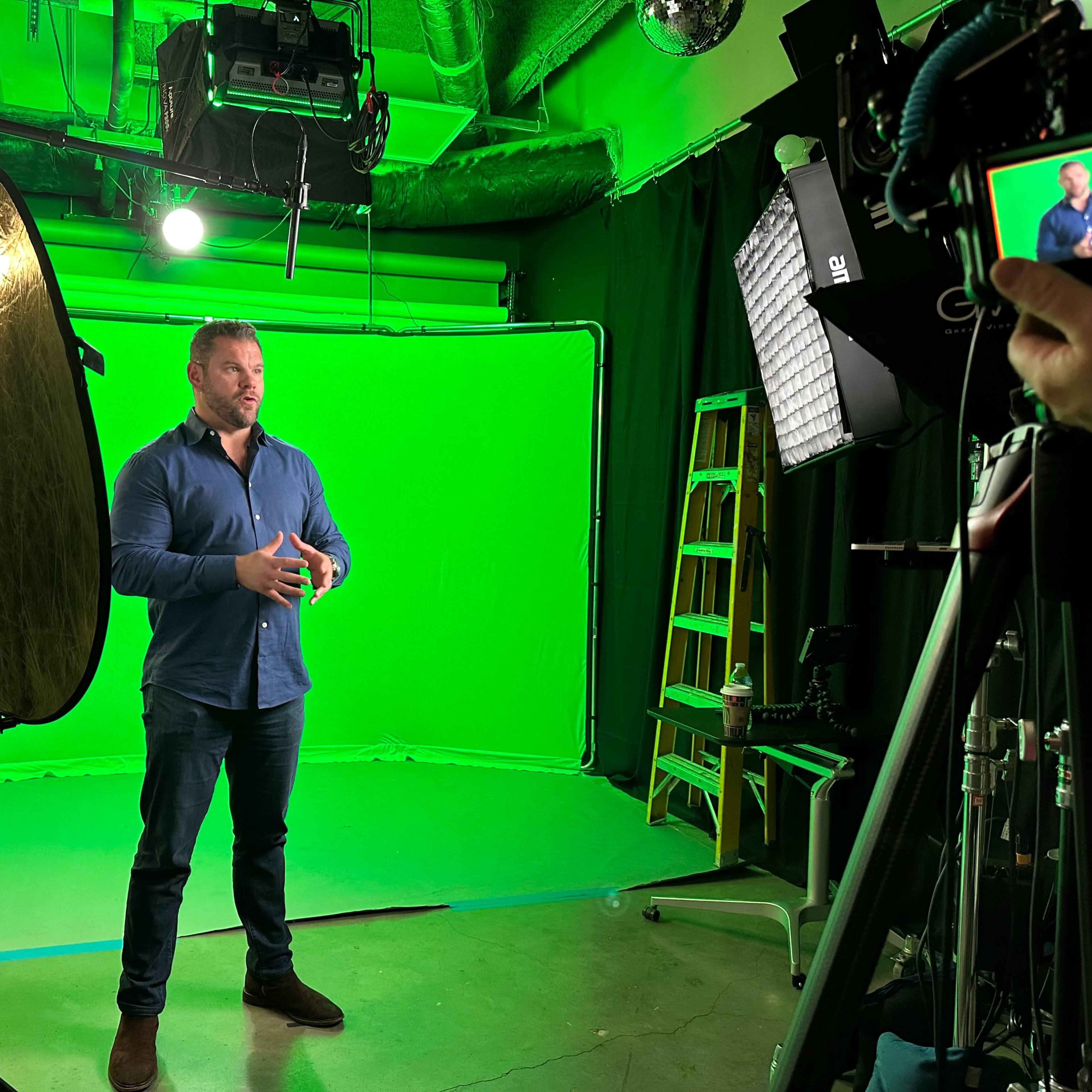 A man stands in front of a green screen, surrounded by various studio lighting equipment and a camera setup.