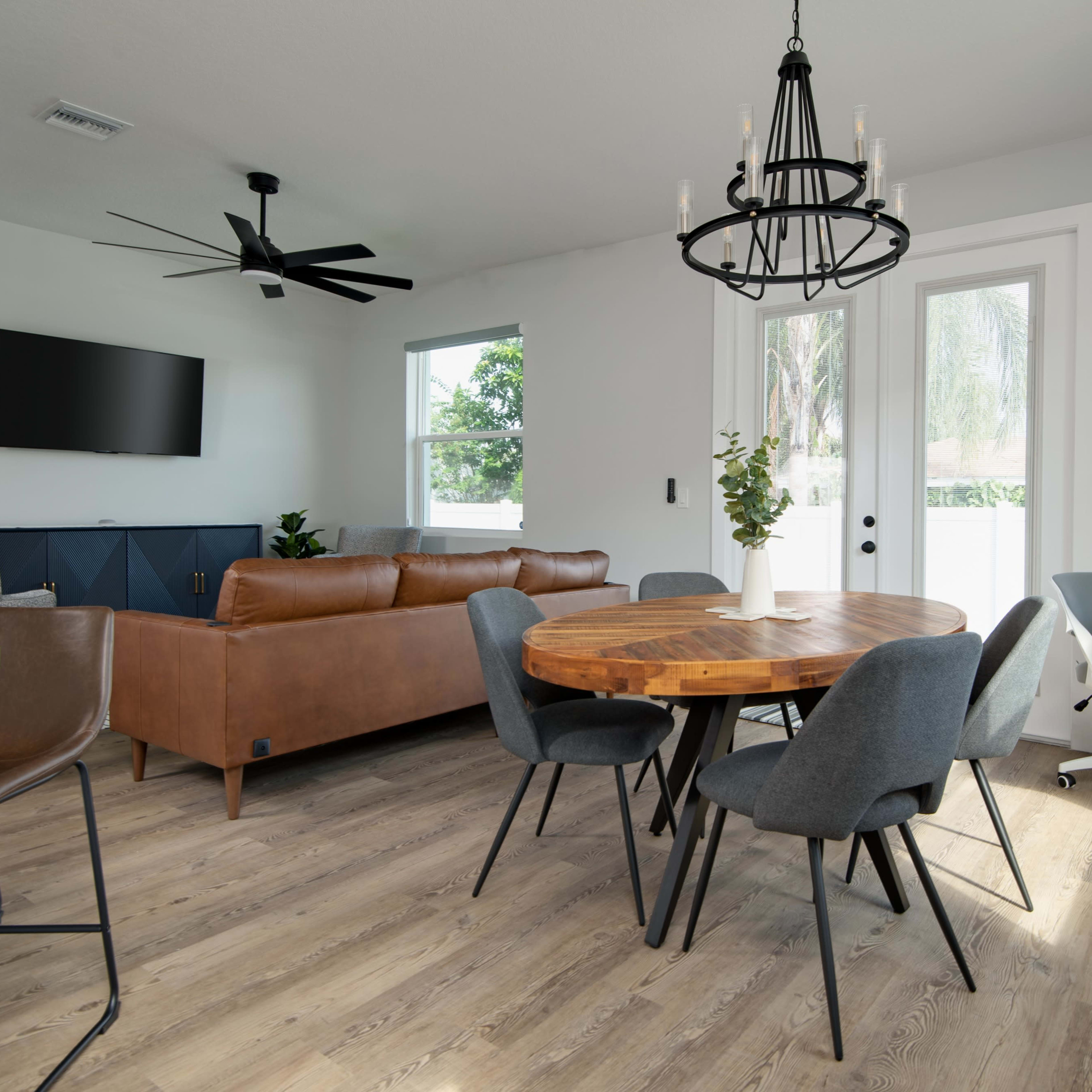A modern living and dining area featuring a round wooden table surrounded by gray chairs, a brown leather sofa, and a large wall-mounted television.