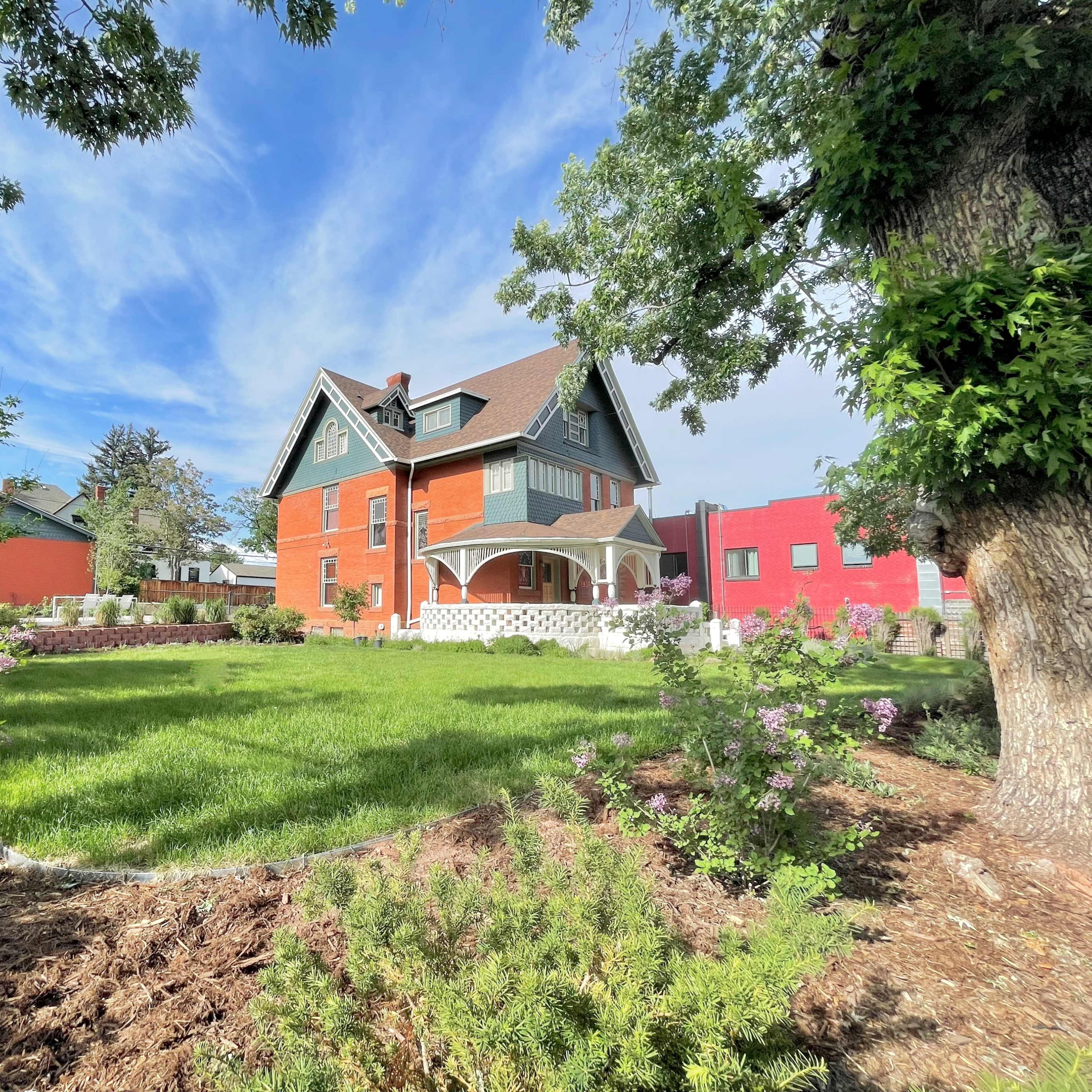 A large, two-story, red and green house sits in a spacious yard surrounded by trees and shrubs.