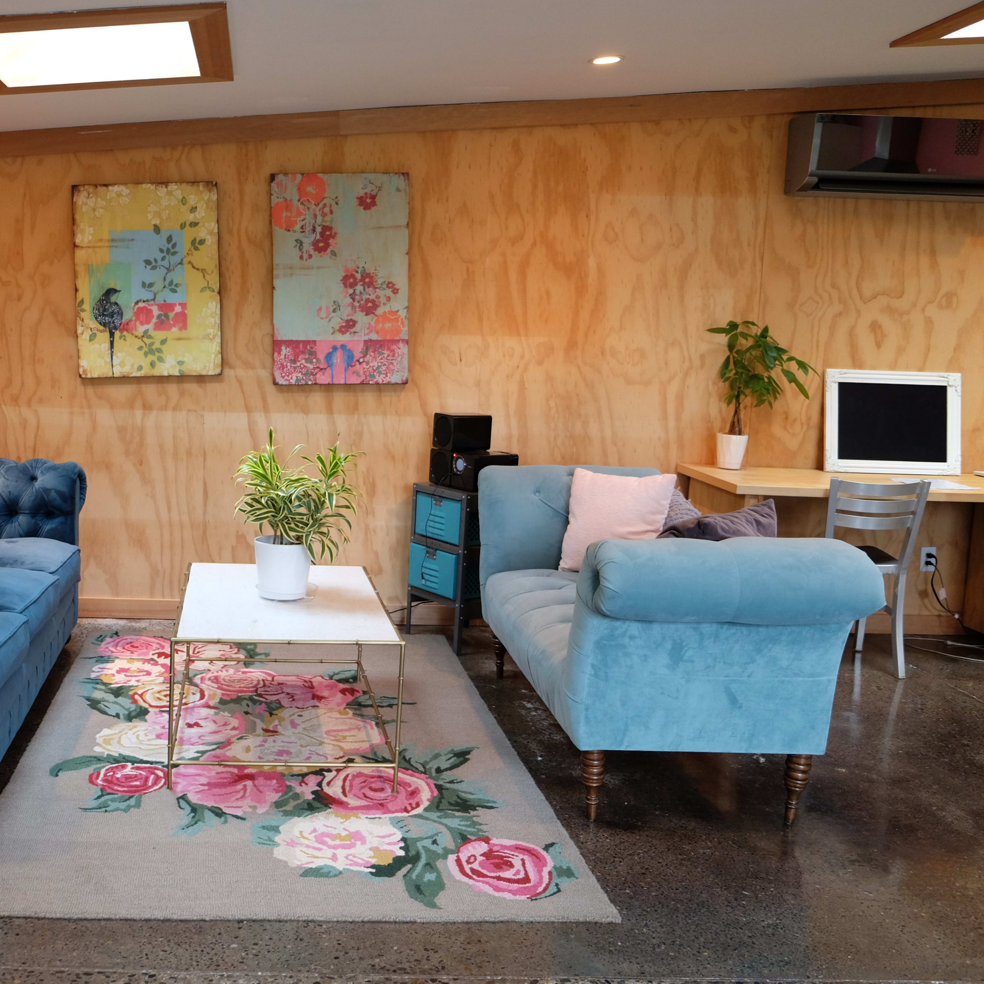 A cozy living area features two blue sofas, a floral rug, a coffee table, and wall art, with wooden paneling and a small desk in the corner.