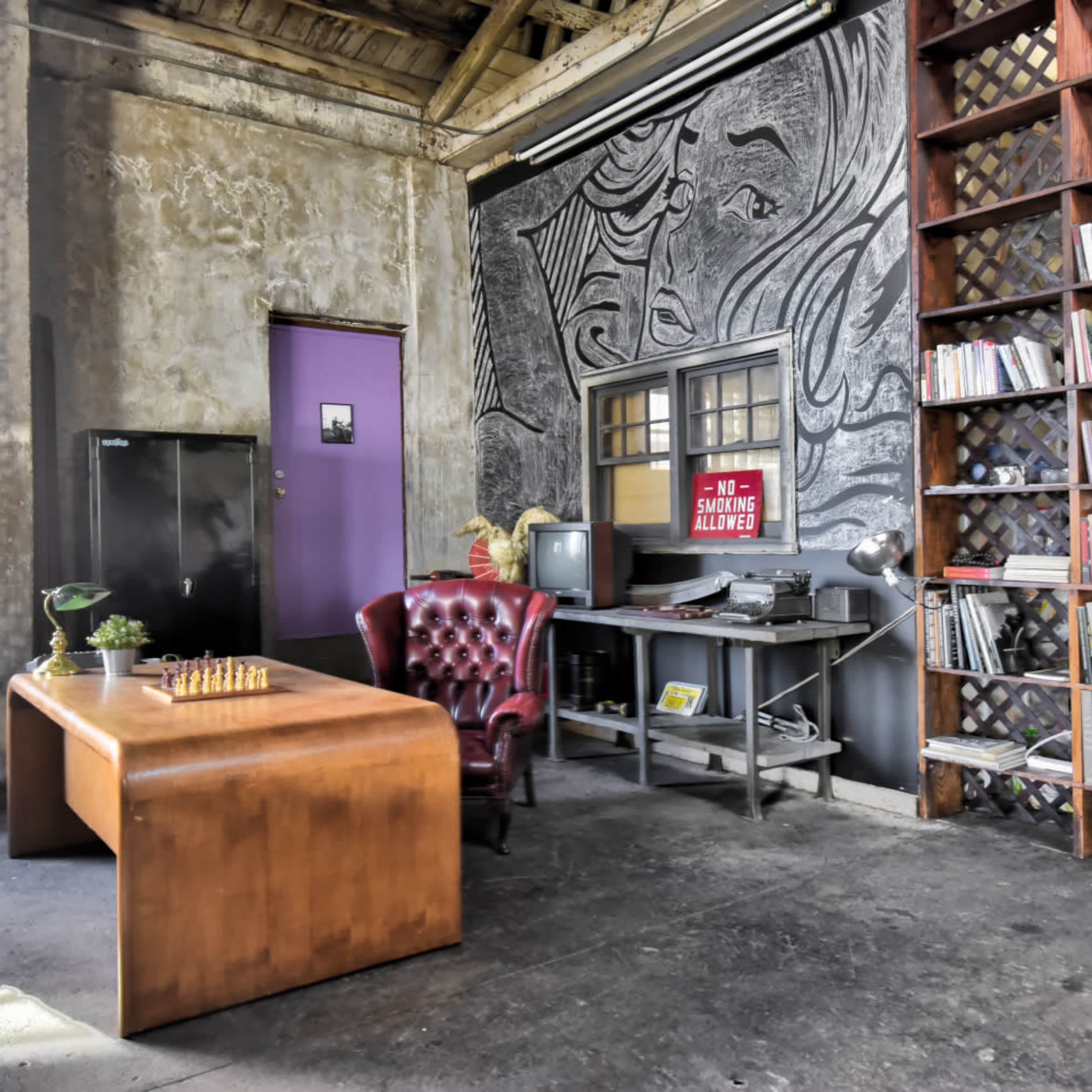 A spacious industrial-style office featuring a wooden desk, a vintage armchair, a bookshelf filled with books, and artwork on the wall.