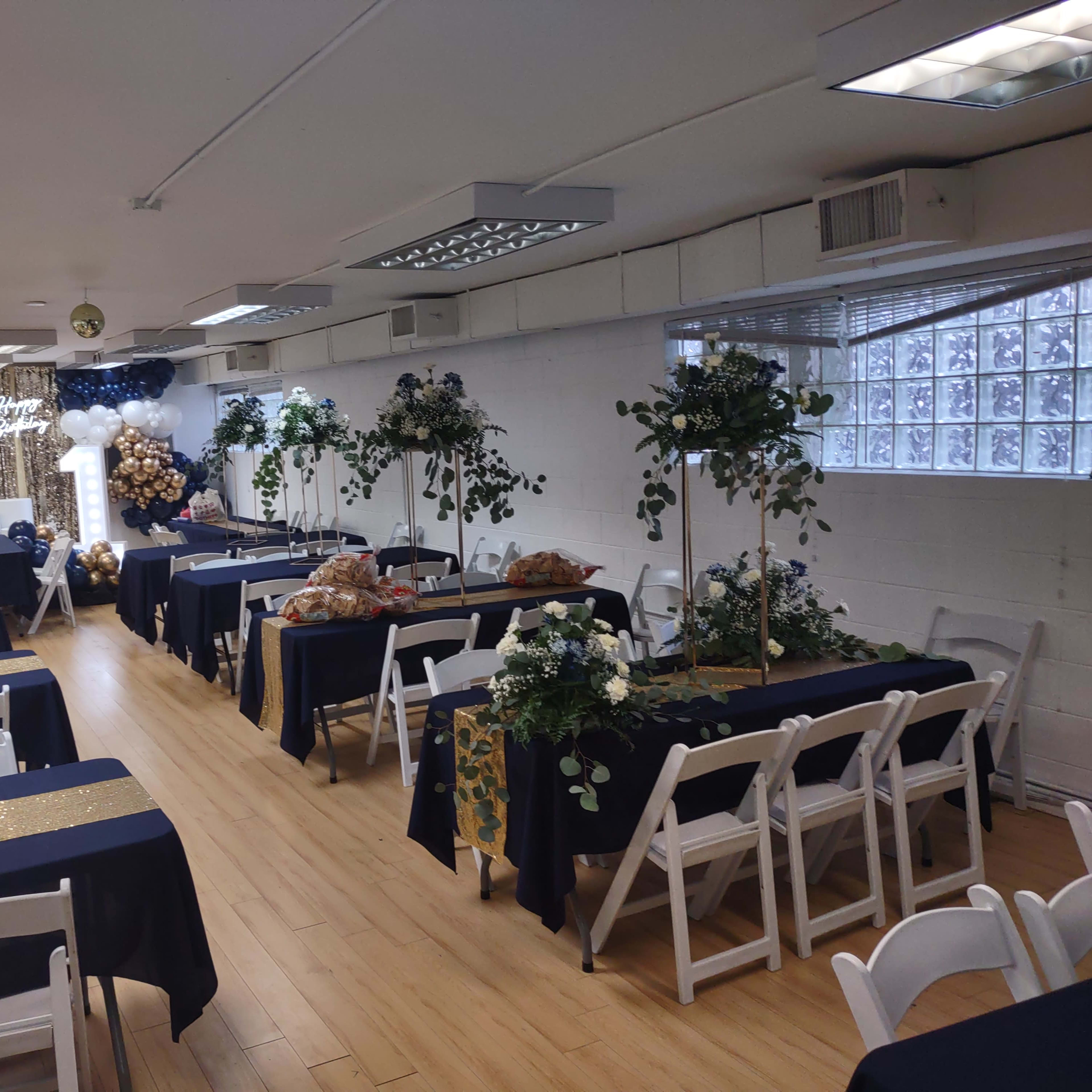 A decorated indoor event space with tables set up for a gathering, featuring blue and gold tablecloths, floral arrangements, and a backdrop with balloons.