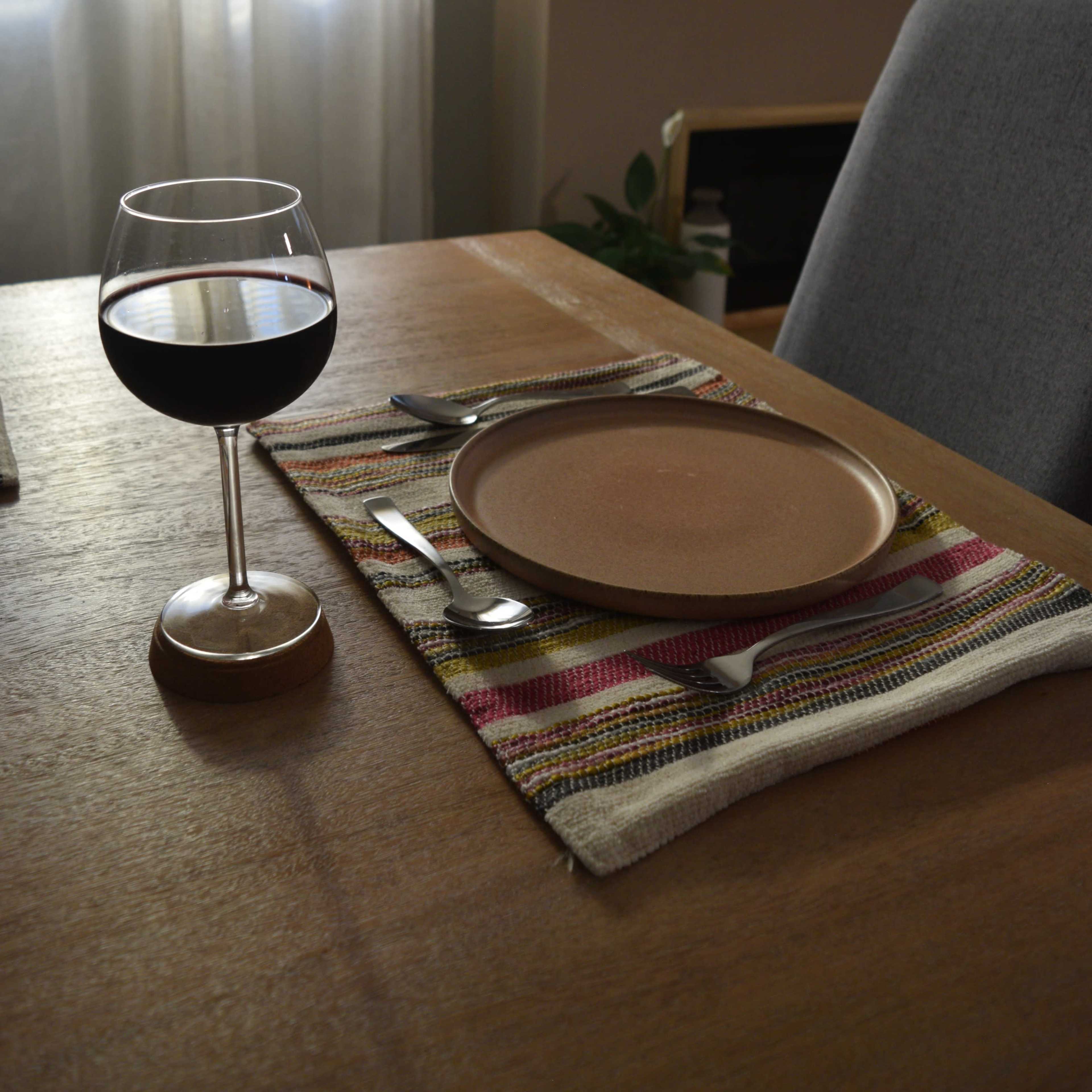 A glass of red wine is placed next to a brown plate on a patterned table mat, set on a wooden table in a softly lit room.