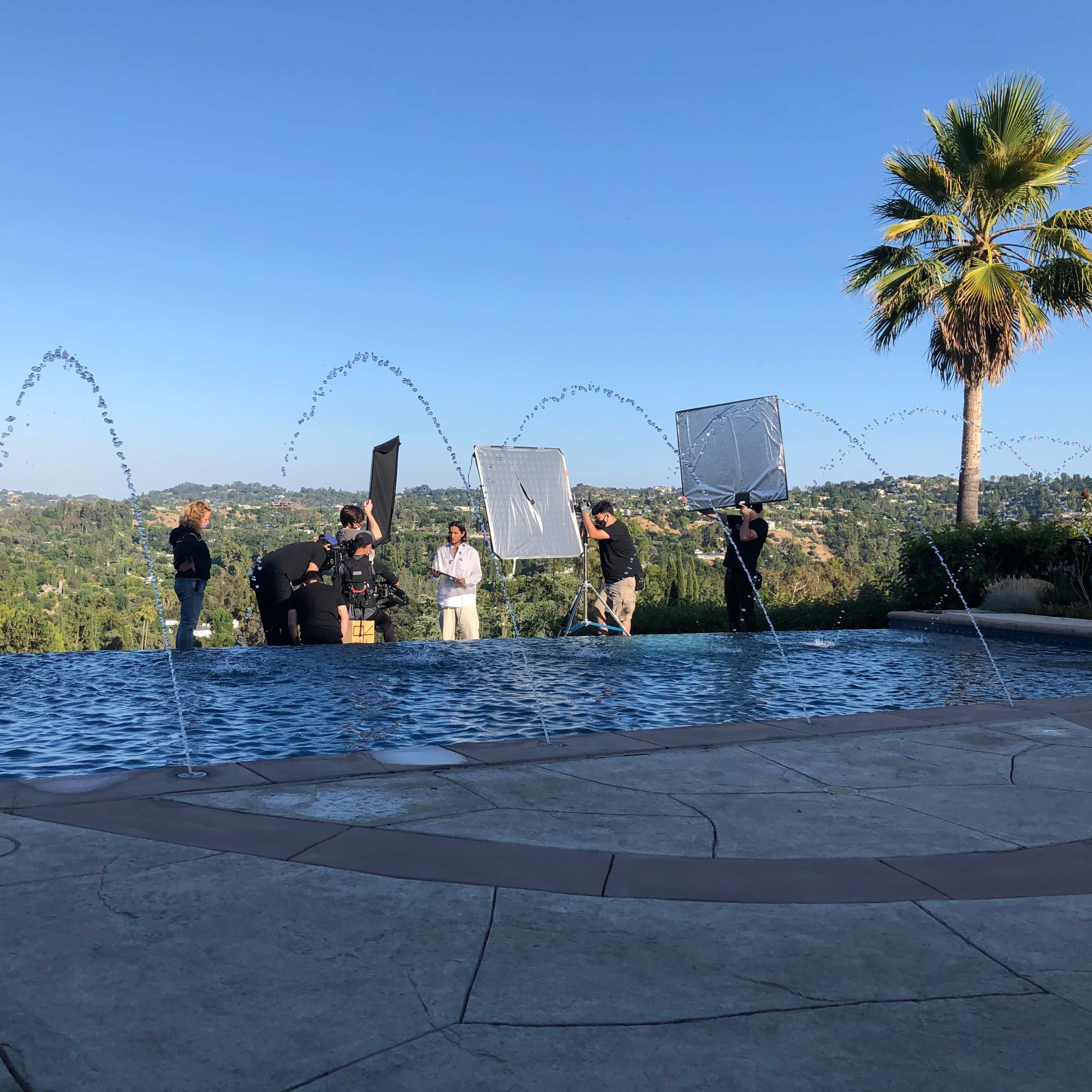 A group of people is working near a pool with fountains, using reflectors and lights against a backdrop of hills and a clear blue sky.