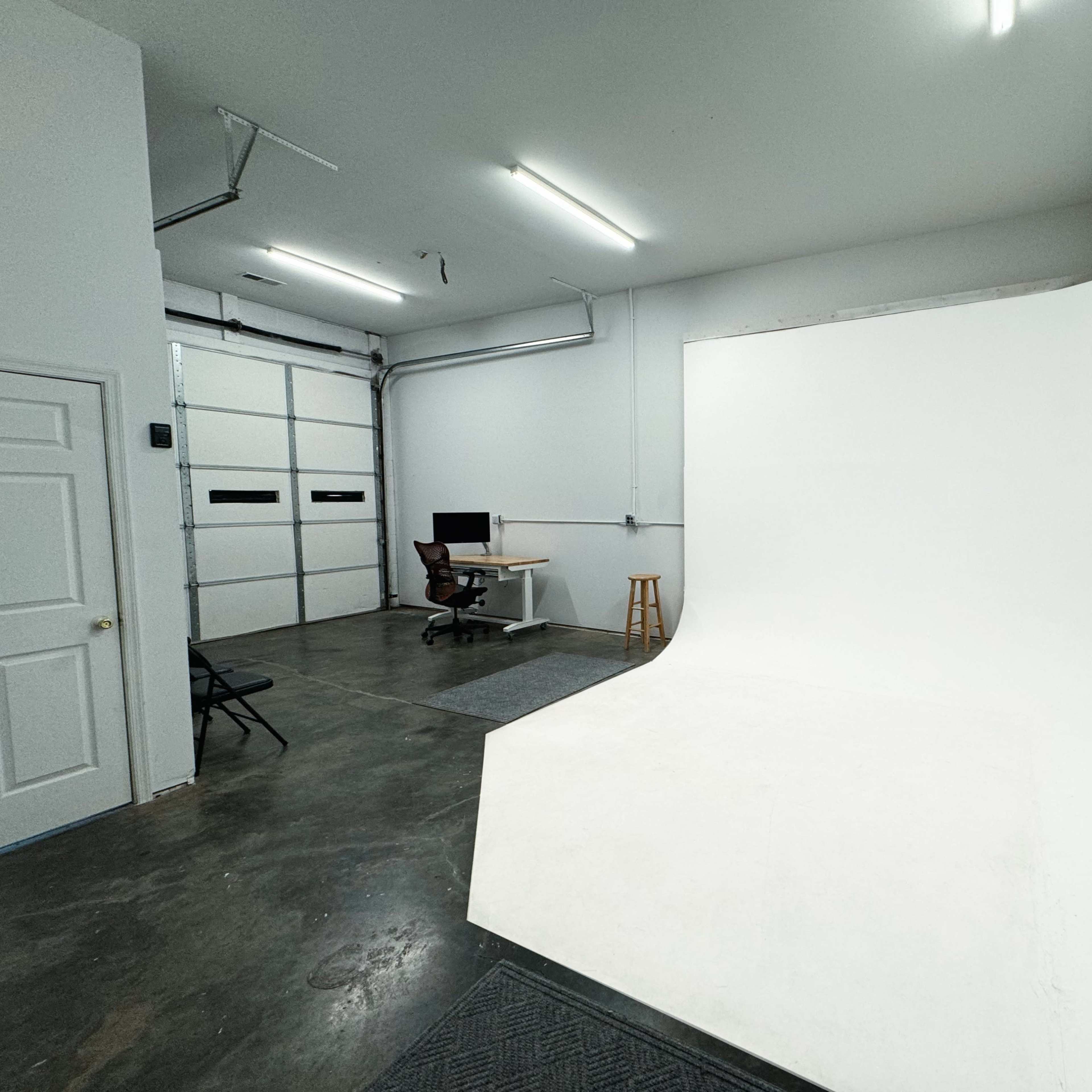 An empty garage space with a seamless white backdrop on one side and a desk with a computer in the corner.