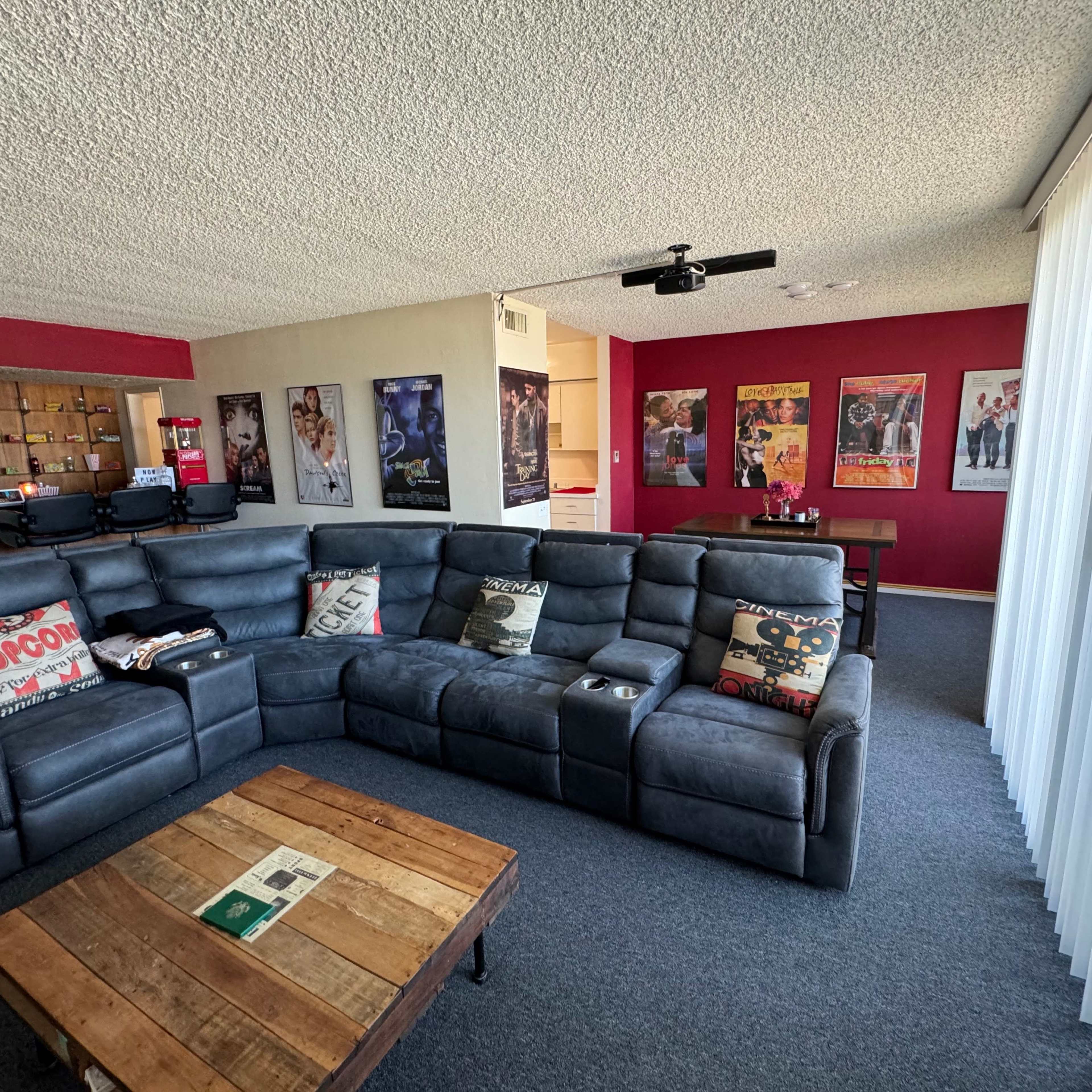A modern living room features a large dark sectional sofa, a wooden coffee table, and walls adorned with movie posters.