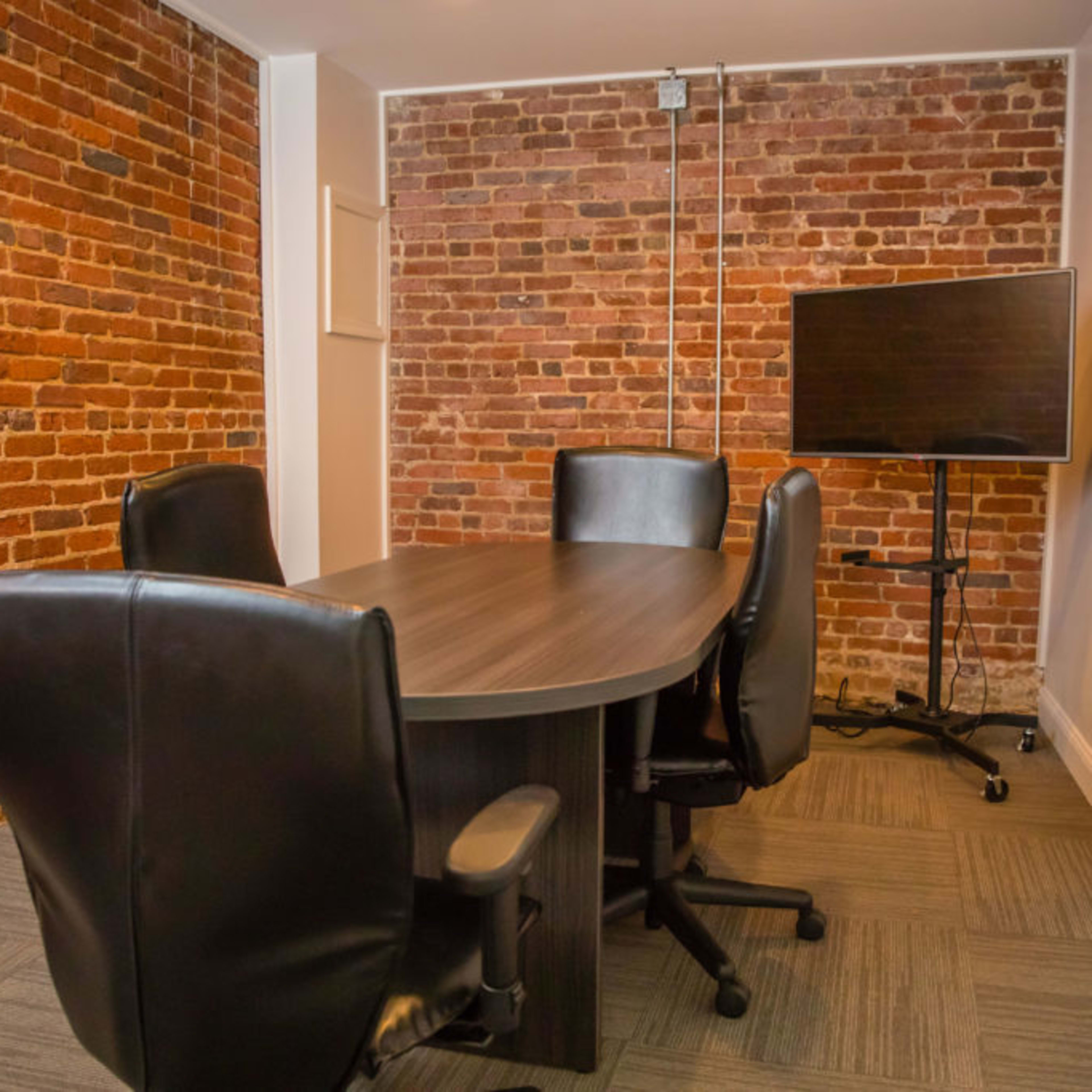 A conference room featuring a wooden table surrounded by four black chairs, with a brick wall and a mounted television.