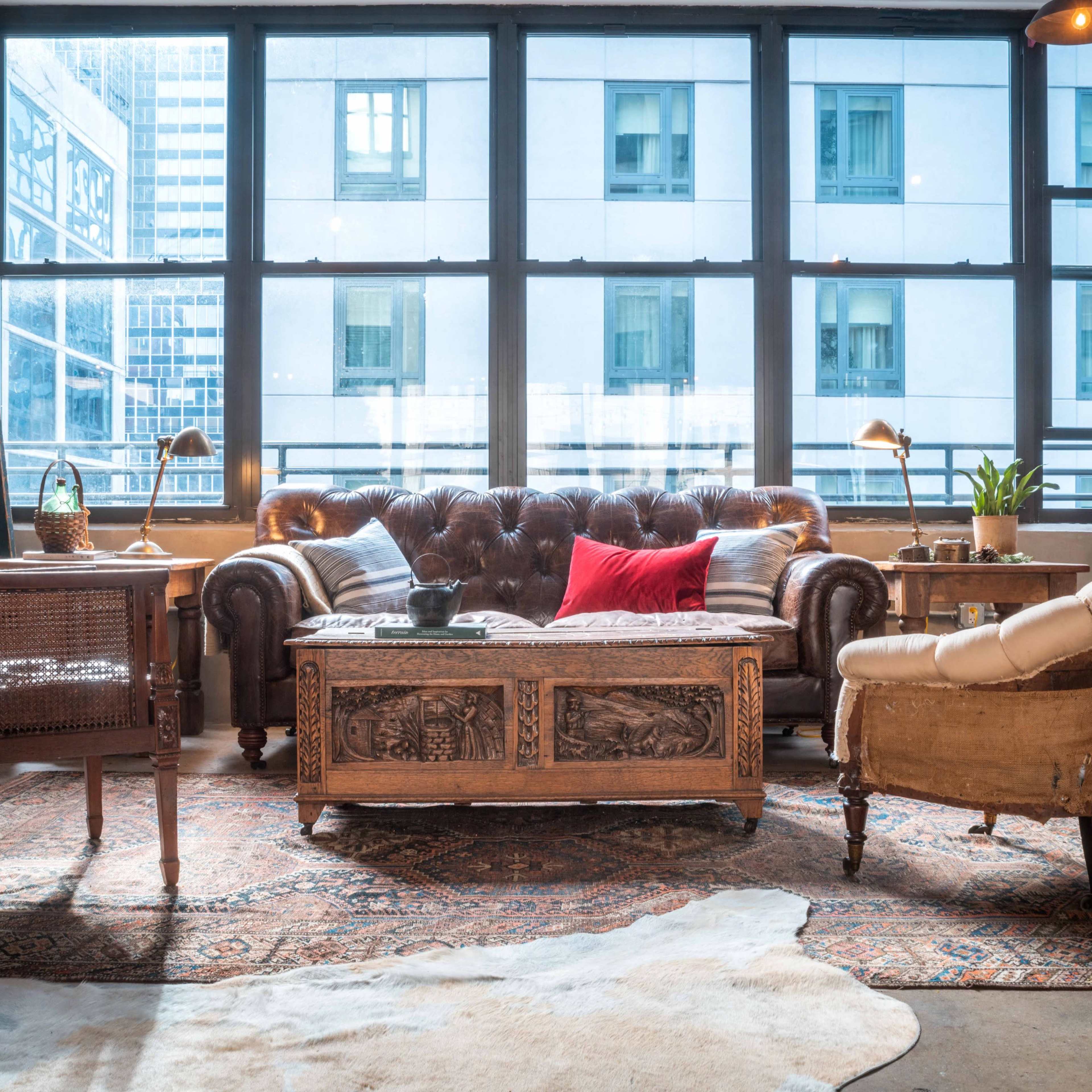 A cozy living area with a brown leather tufted sofa, two chairs, an ornate wooden coffee table, and large windows allowing natural light.