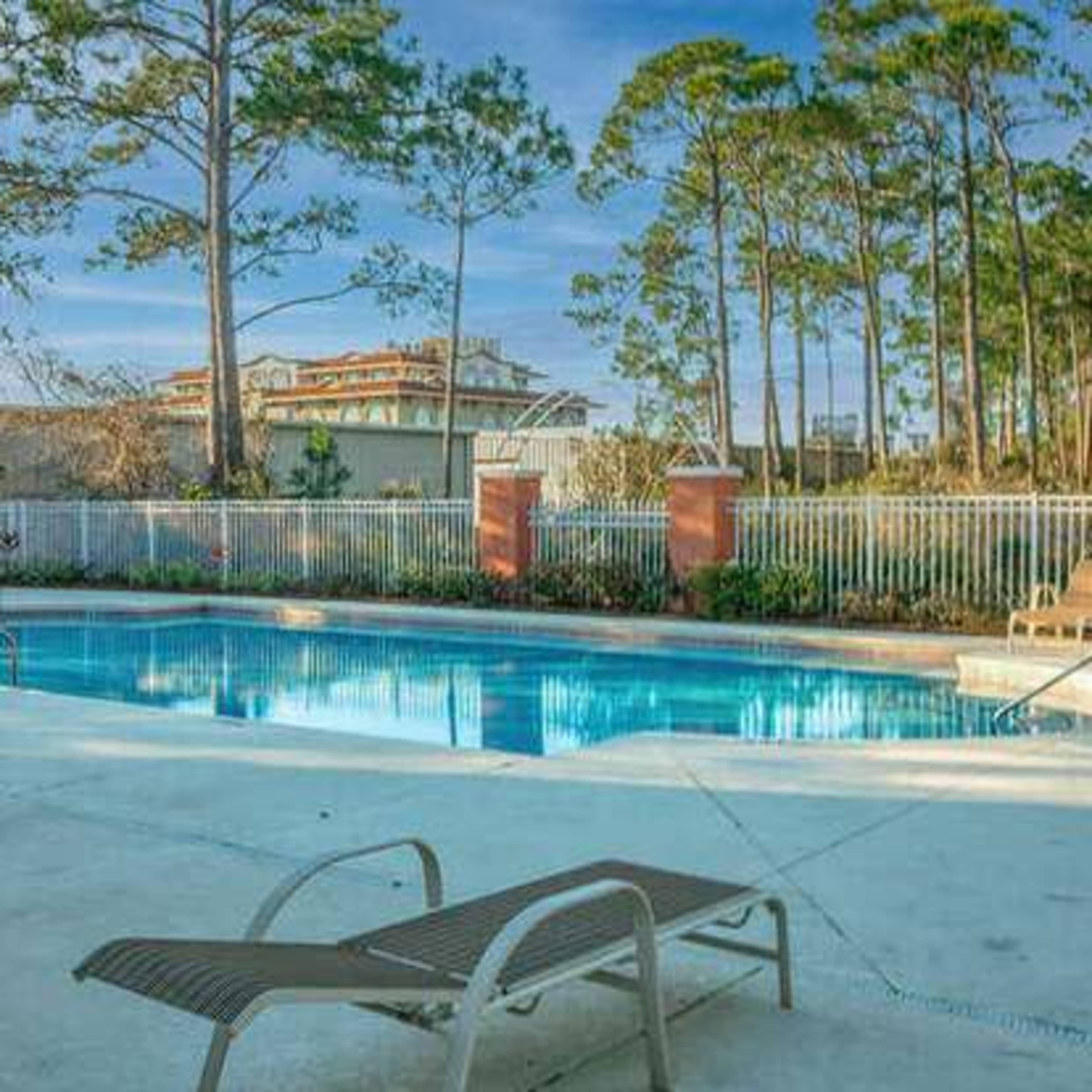 A clear swimming pool surrounded by tan lounge chairs and a fence, with tall trees and buildings in the background.