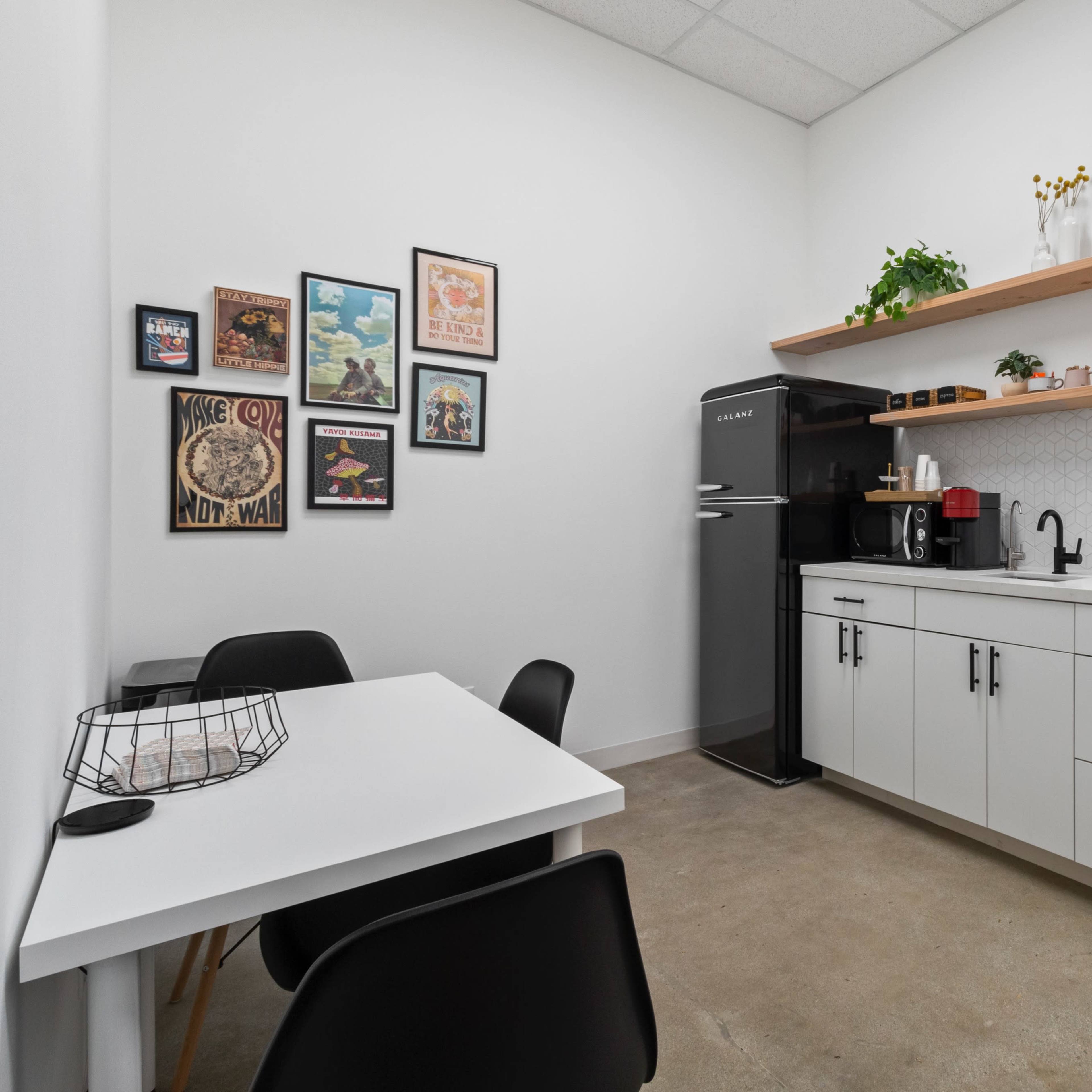 A small kitchen area with a black refrigerator, white cabinets, and a table with two chairs, decorated with wall art and a few potted plants.