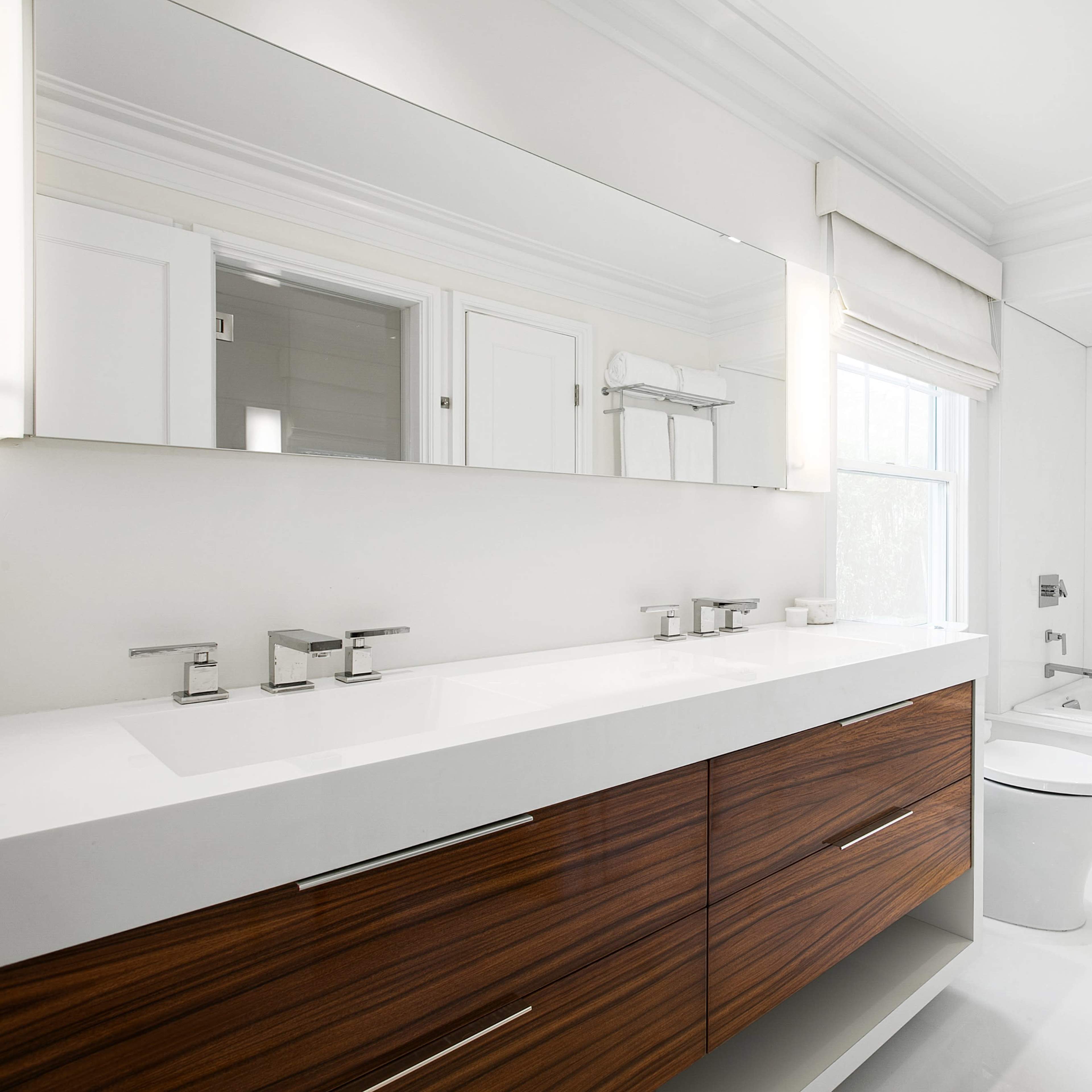 The bathroom features a double sink vanity with wooden cabinetry, a large mirror, a freestanding bathtub, and a white toilet against a bright, neutral background.