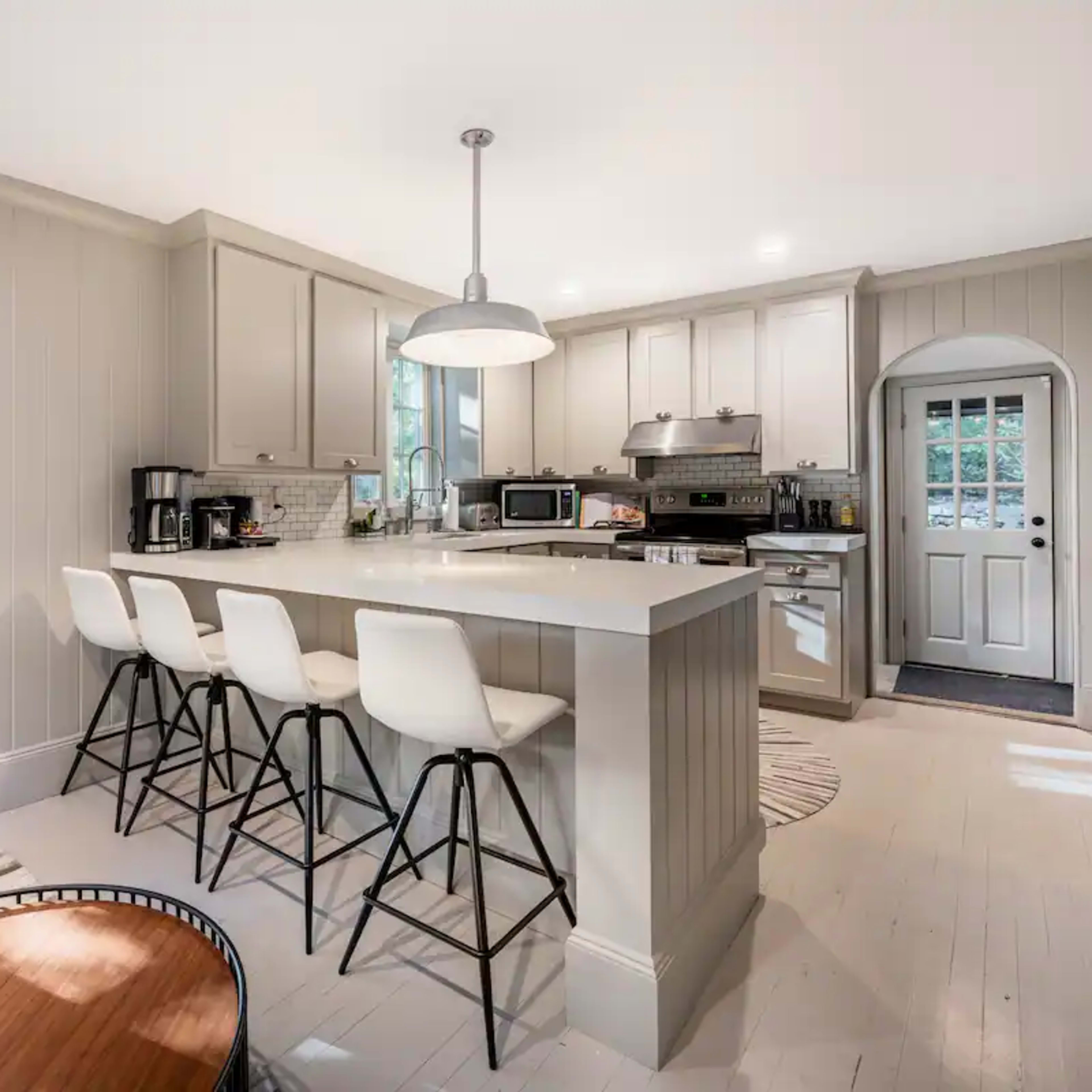 A modern kitchen features a central island with bar stools, stainless steel appliances, and windows allowing natural light.