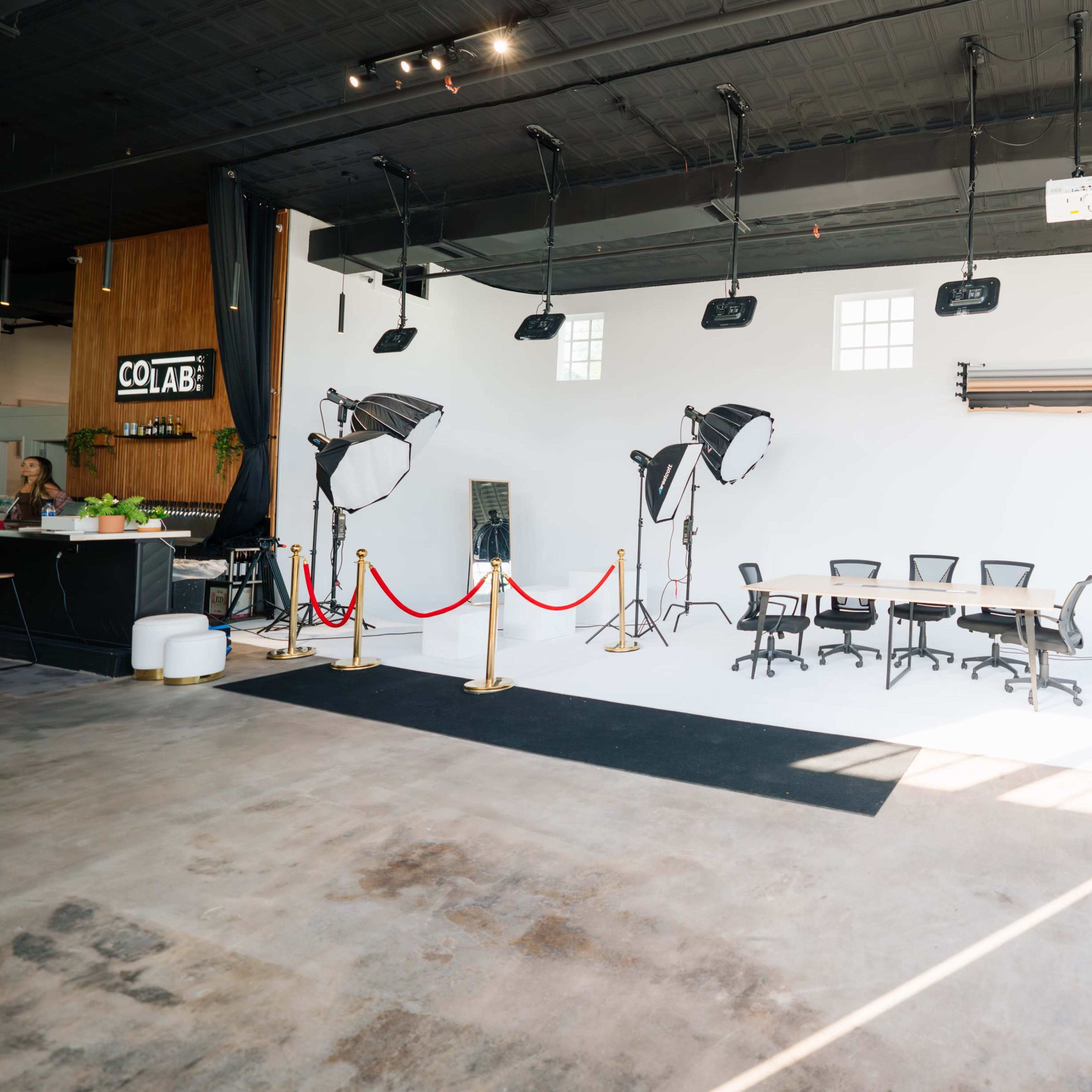 A spacious studio with a large white backdrop, several lights, and a round table surrounded by office chairs.