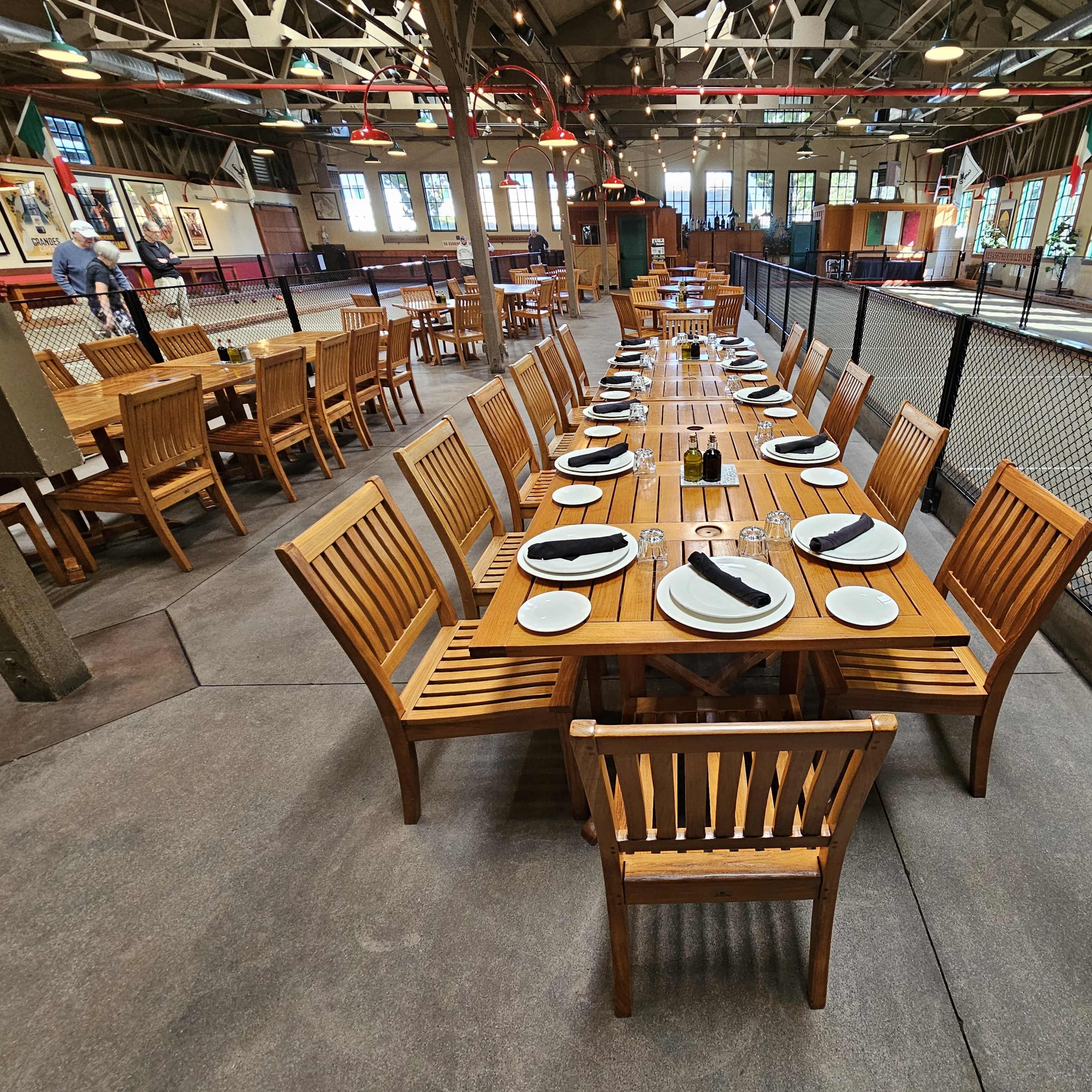 A long wooden dining table is neatly arranged with plates and napkins in a spacious indoor restaurant setting with large windows.