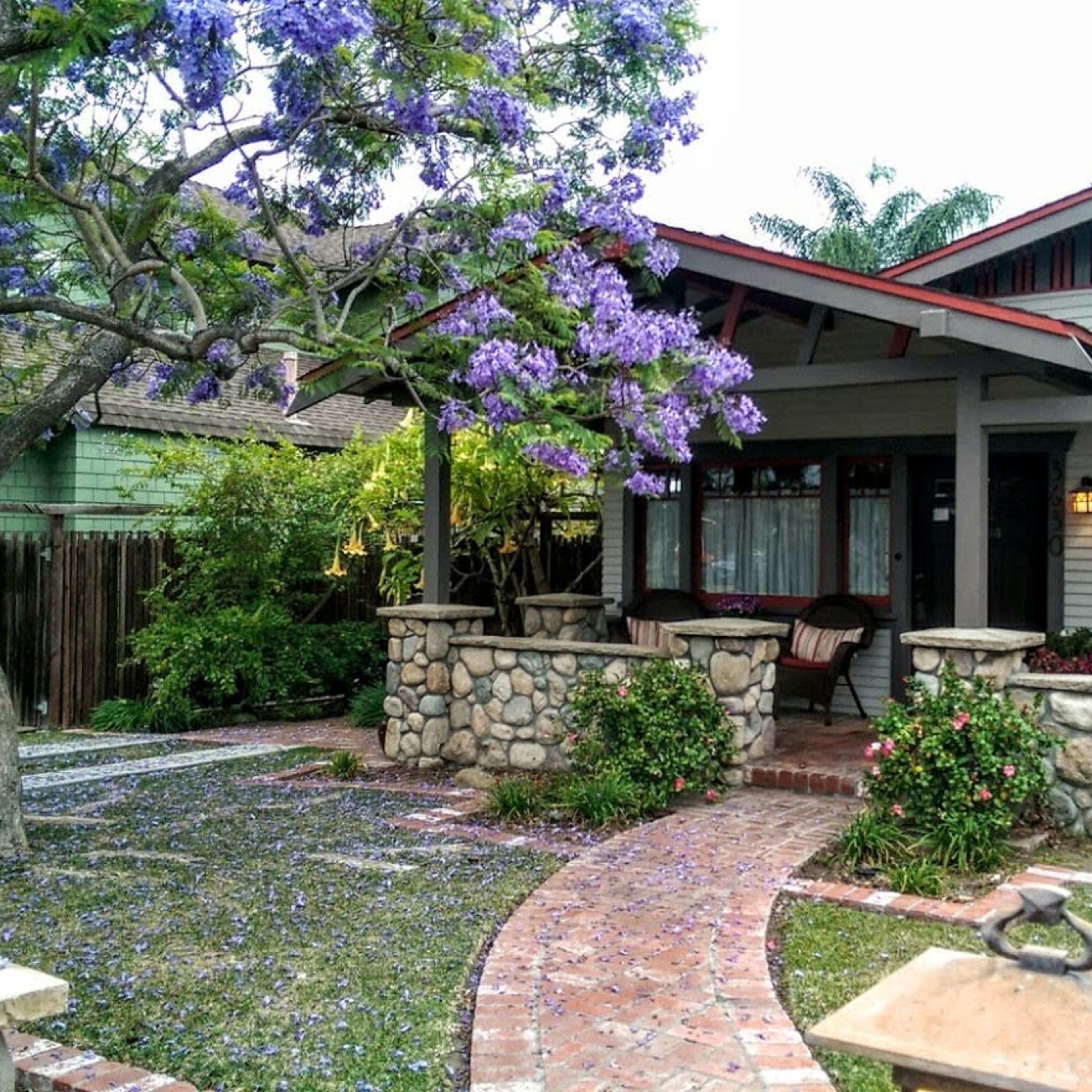 A charming bungalow features a stone pathway leading to its front porch, surrounded by a landscaped yard with purple flowering trees.