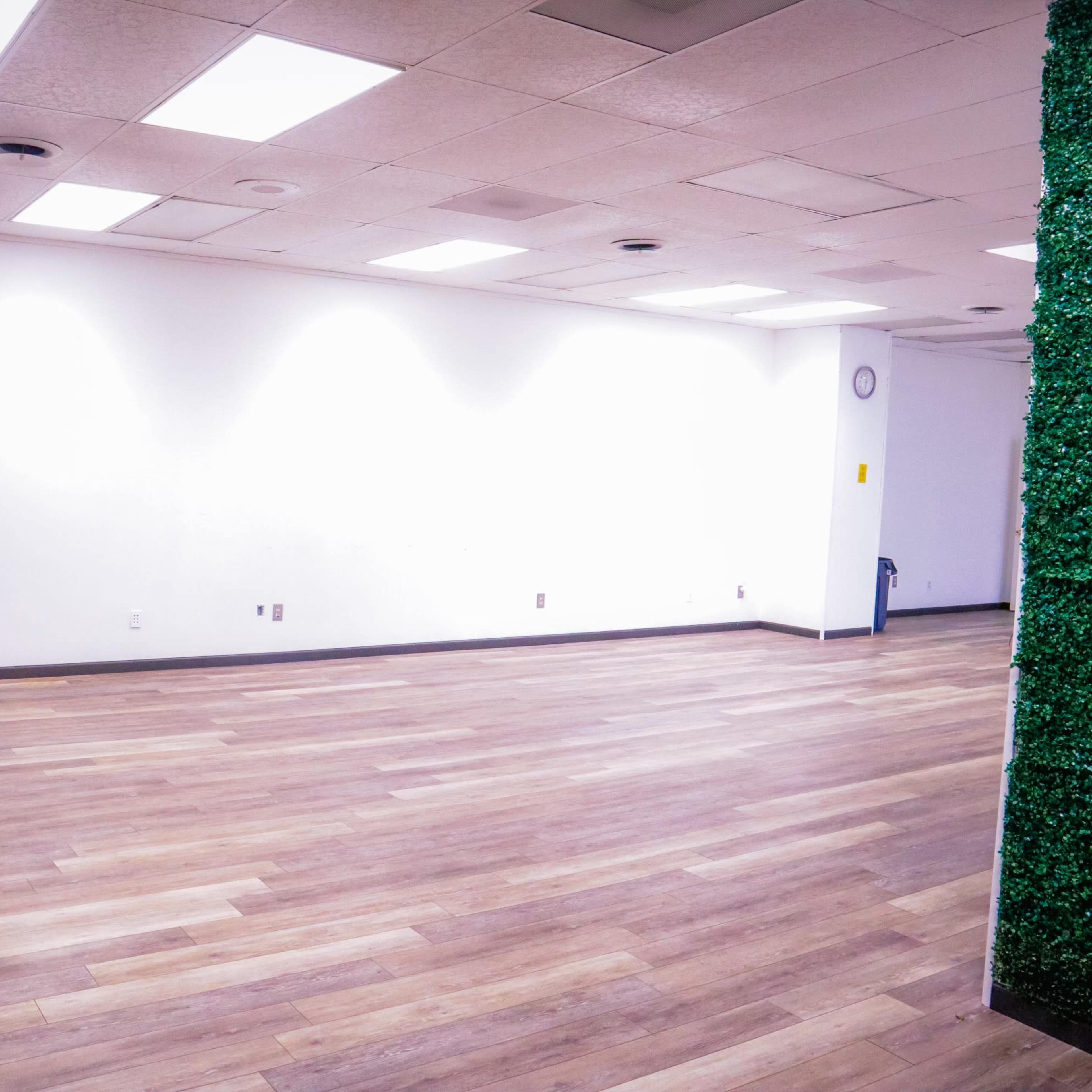 An empty room with hardwood floors, featuring a gray armchair and a small area covered in green plants on one side.