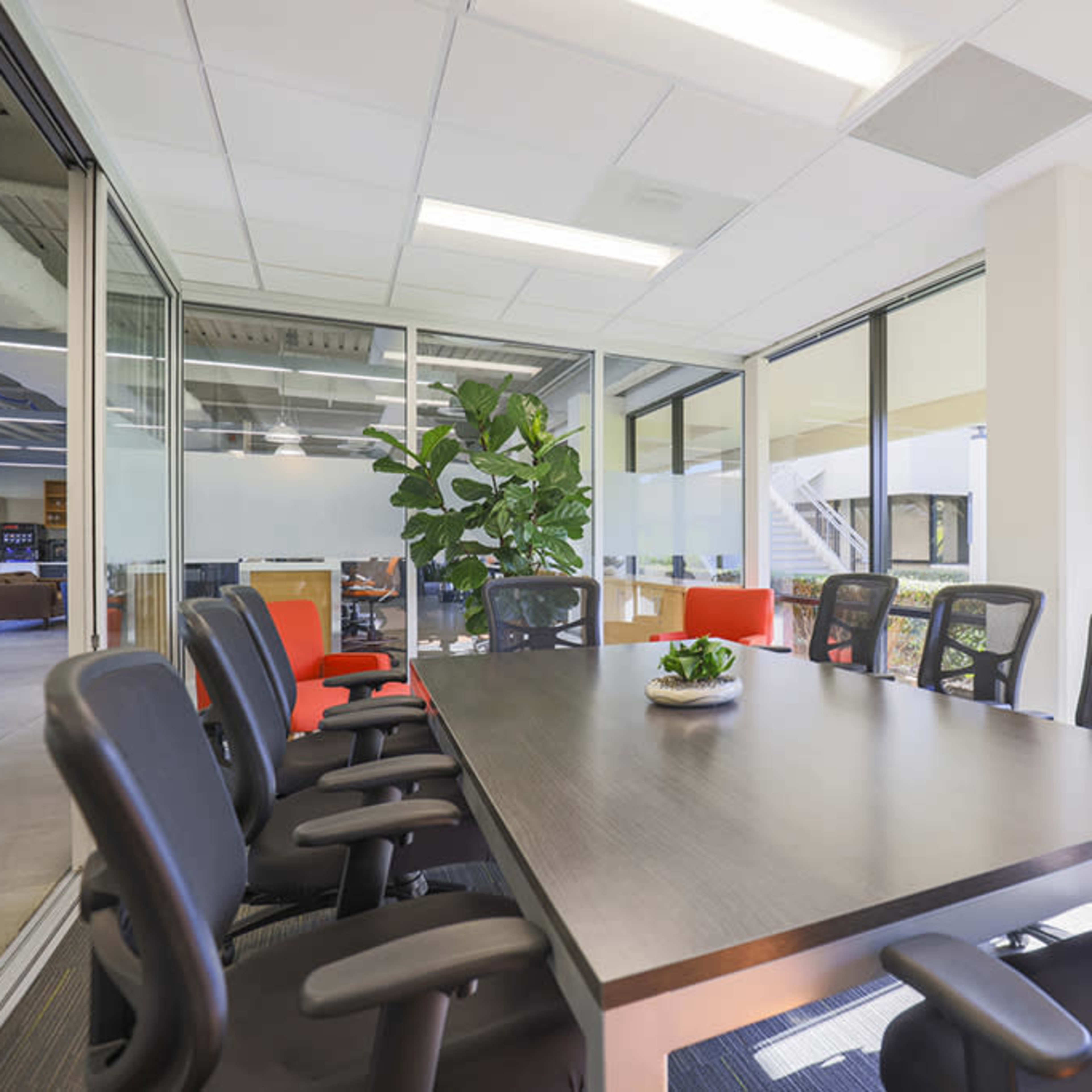 A modern conference room features a long table surrounded by black chairs, with large windows allowing natural light and a view of greenery outside.