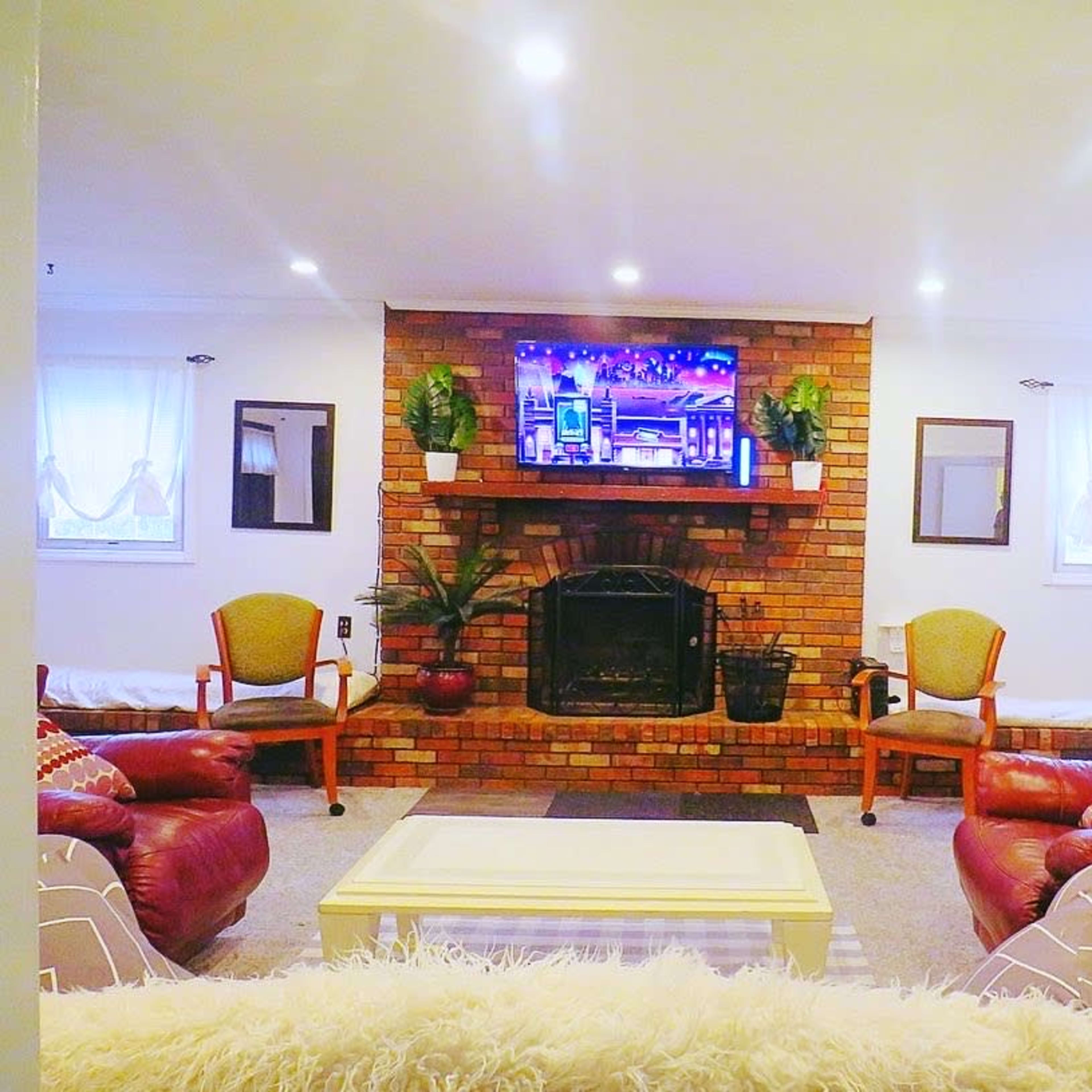 A living room with a brick fireplace, a large television mounted above it, and various seating arrangements including two chairs and a sofa.