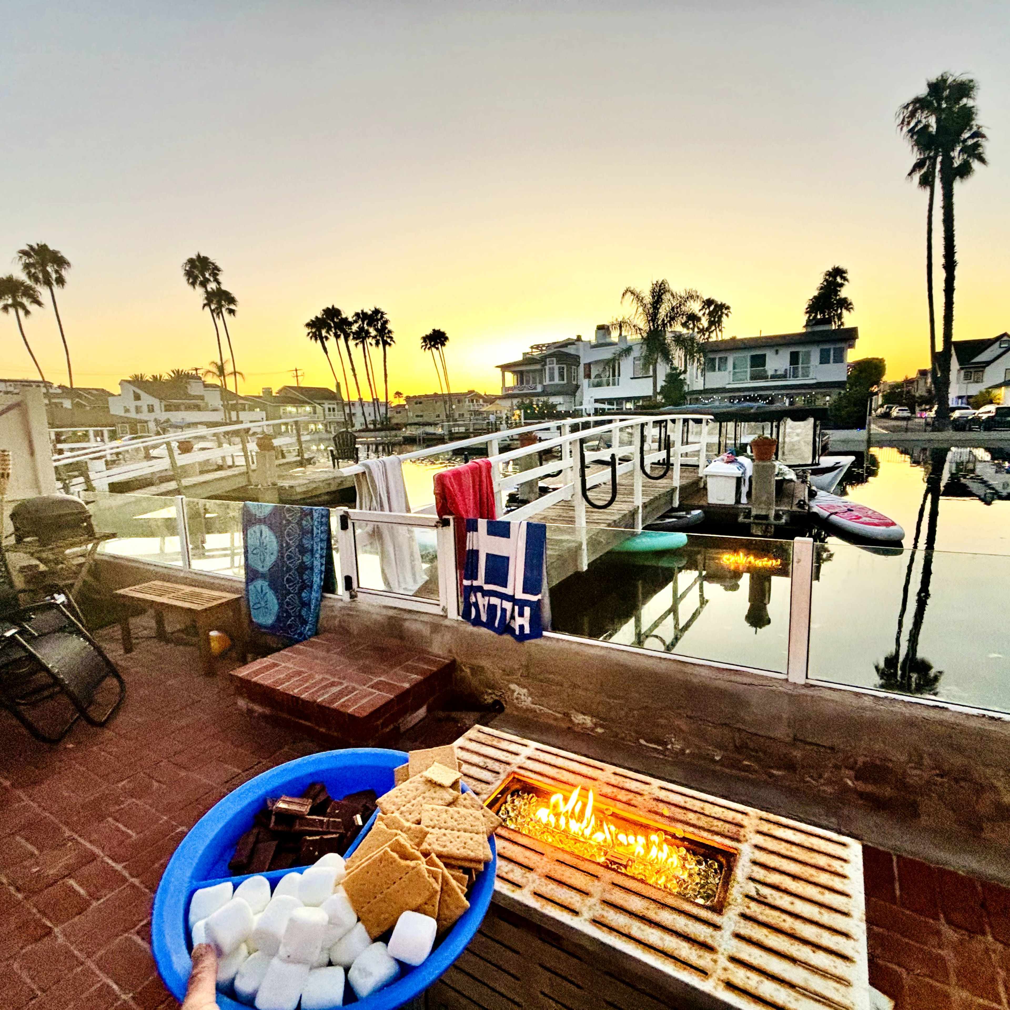 A person holds a blue bowl of s'mores ingredients while overlooking a calm waterfront scene at sunset, with palm trees and houses reflected in the water.
