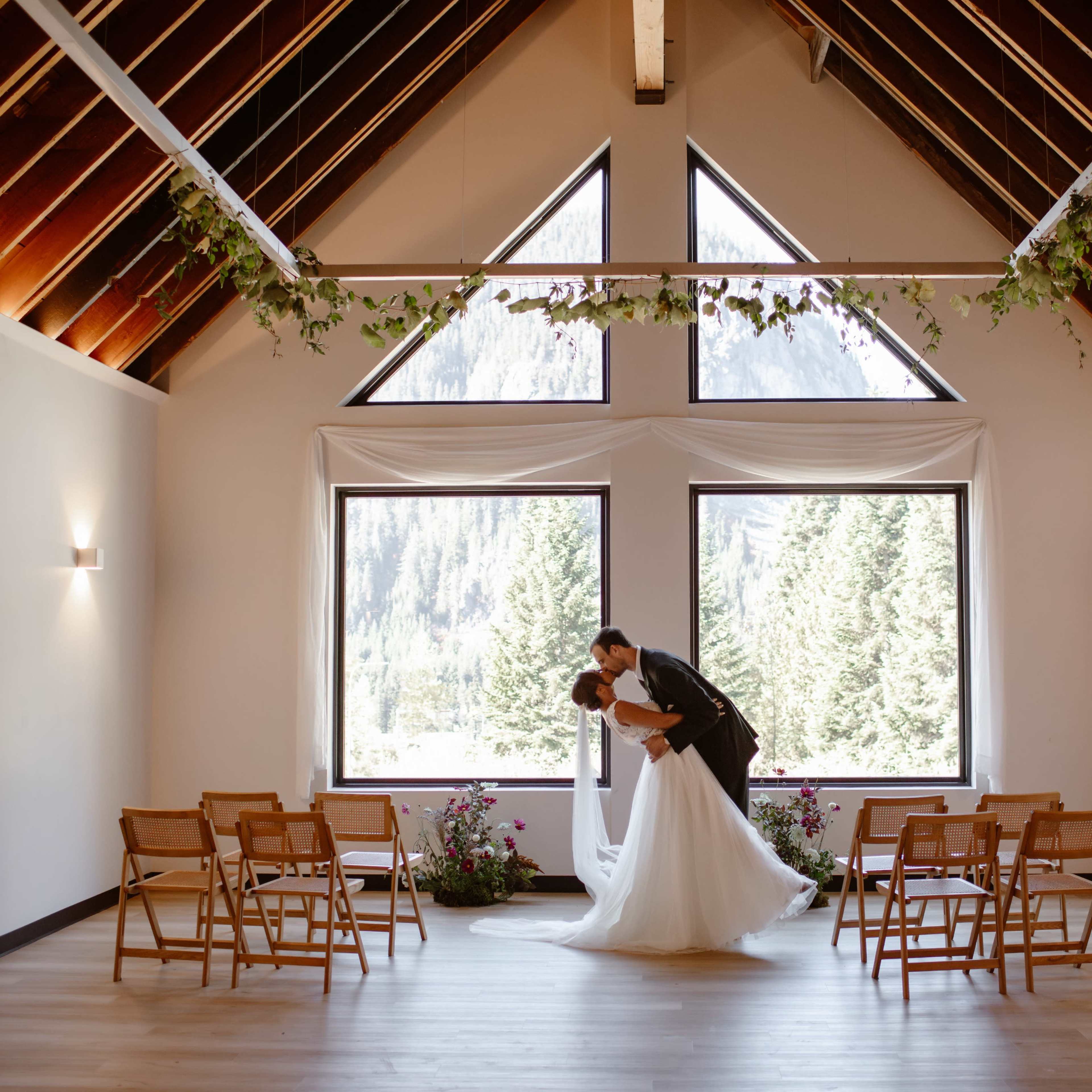 A bride and groom kiss in a spacious, well-lit interior with large windows and wooden accents, surrounded by folding chairs and greenery.