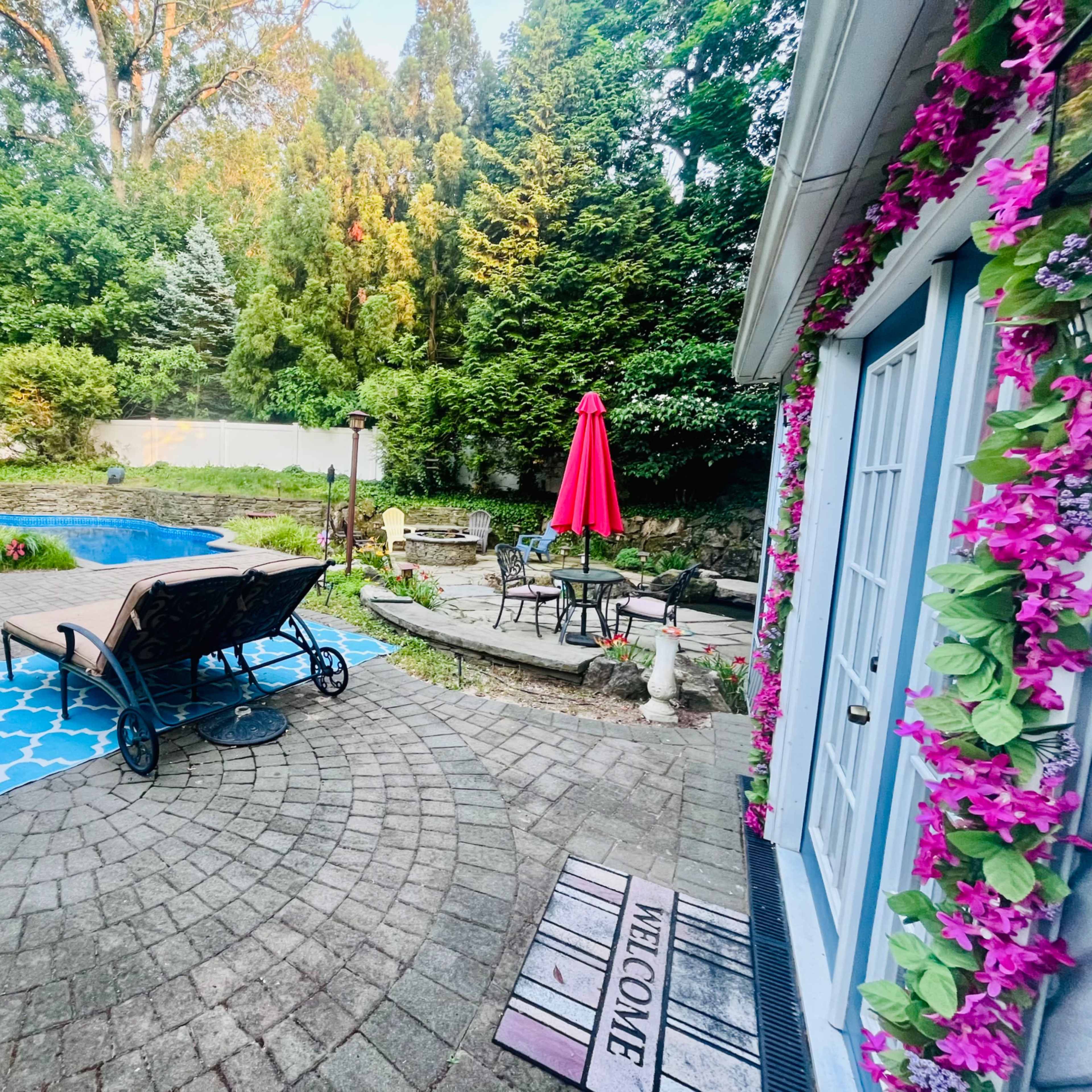 A landscaped backyard with a pool, a lounge chair, a red umbrella, and a patterned rug near a stone pathway.