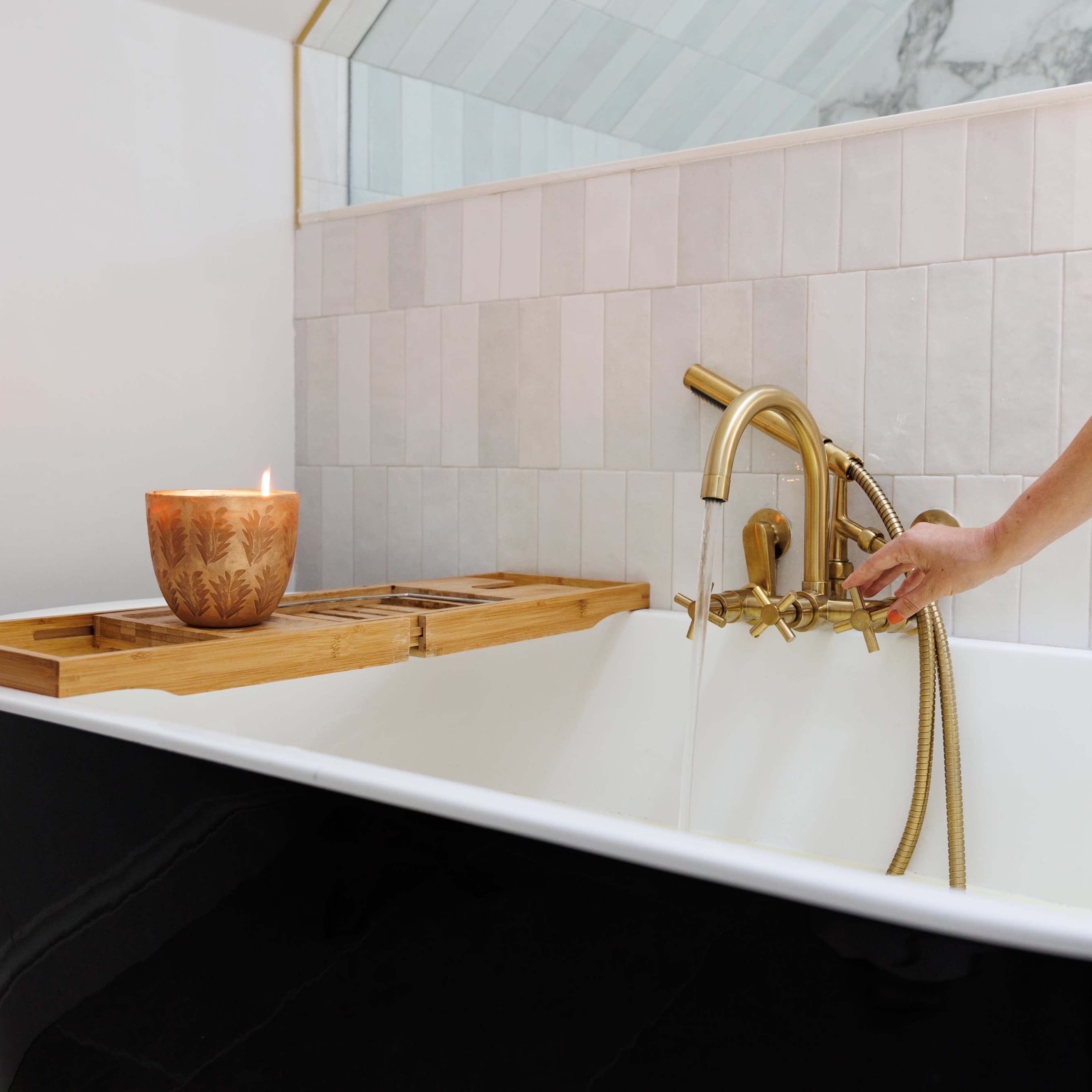 A hand is turning on a brass faucet above a soaking tub, with a lit candle and wooden bath tray beside it.