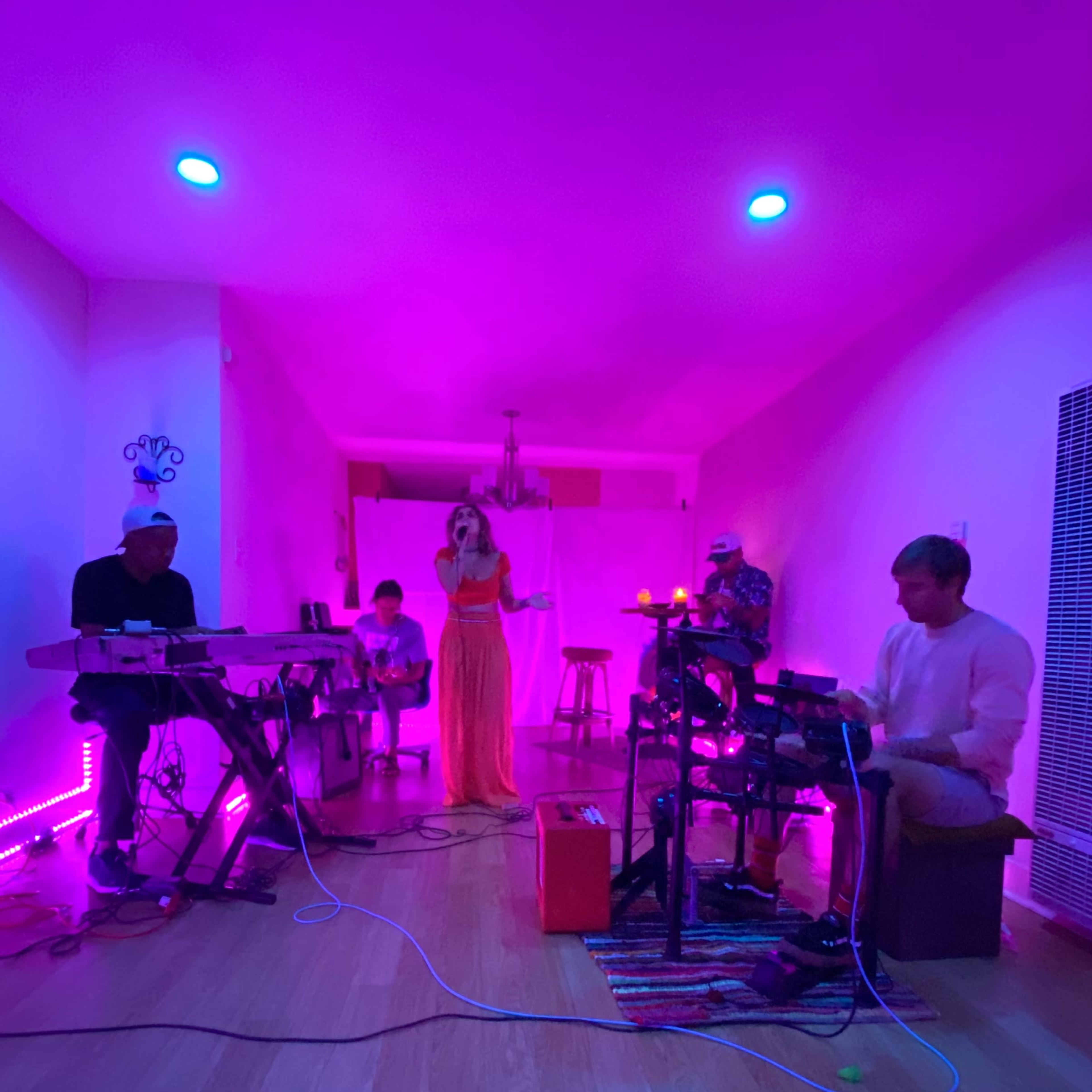 A group of five musicians performs in a dimly lit room with purple lighting, while one singer stands at the front.