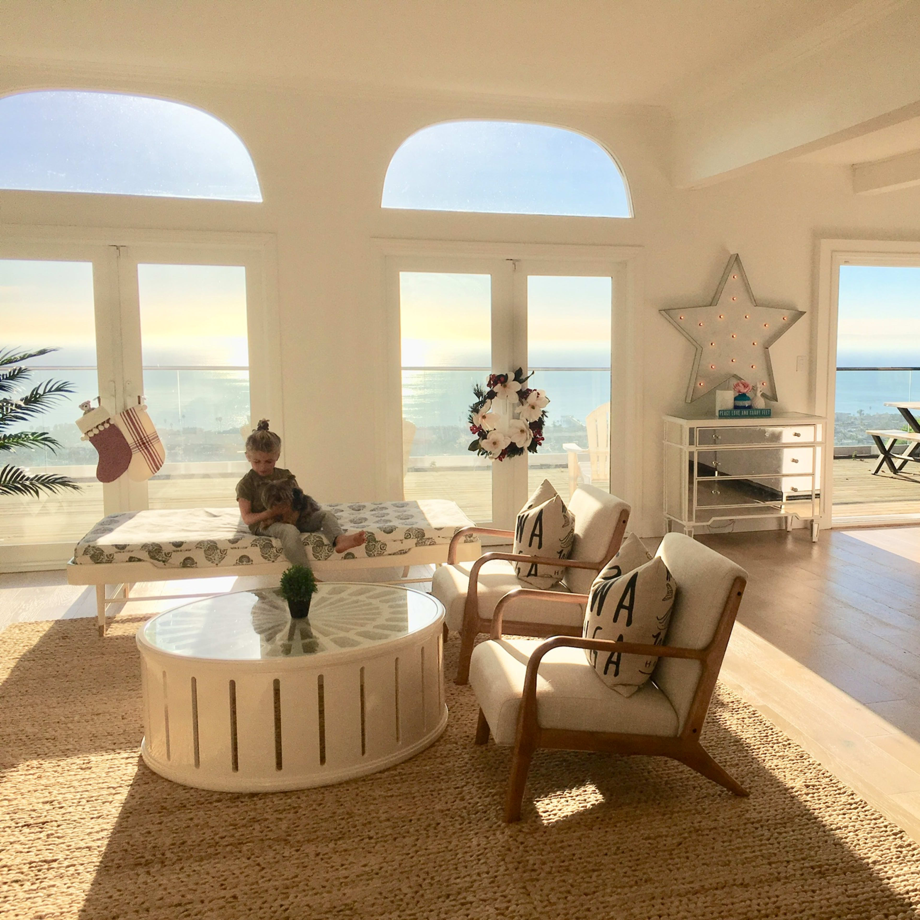 A bright, airy living room features a child sitting on a bench beside a circular coffee table, with large windows showcasing a view of the ocean.