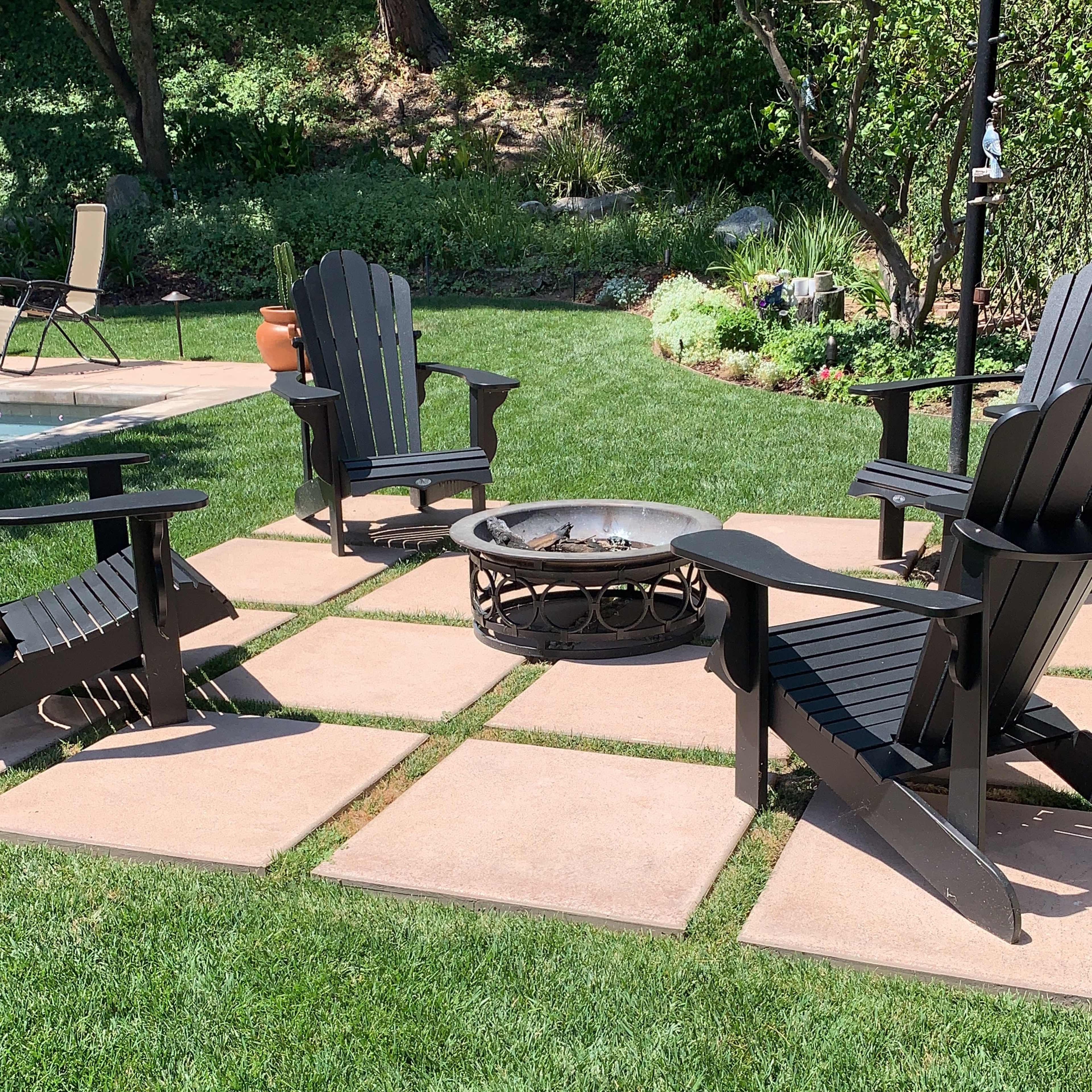 A patio area with four black Adirondack chairs arranged around a stone fire pit, surrounded by well-maintained grass and landscaping.