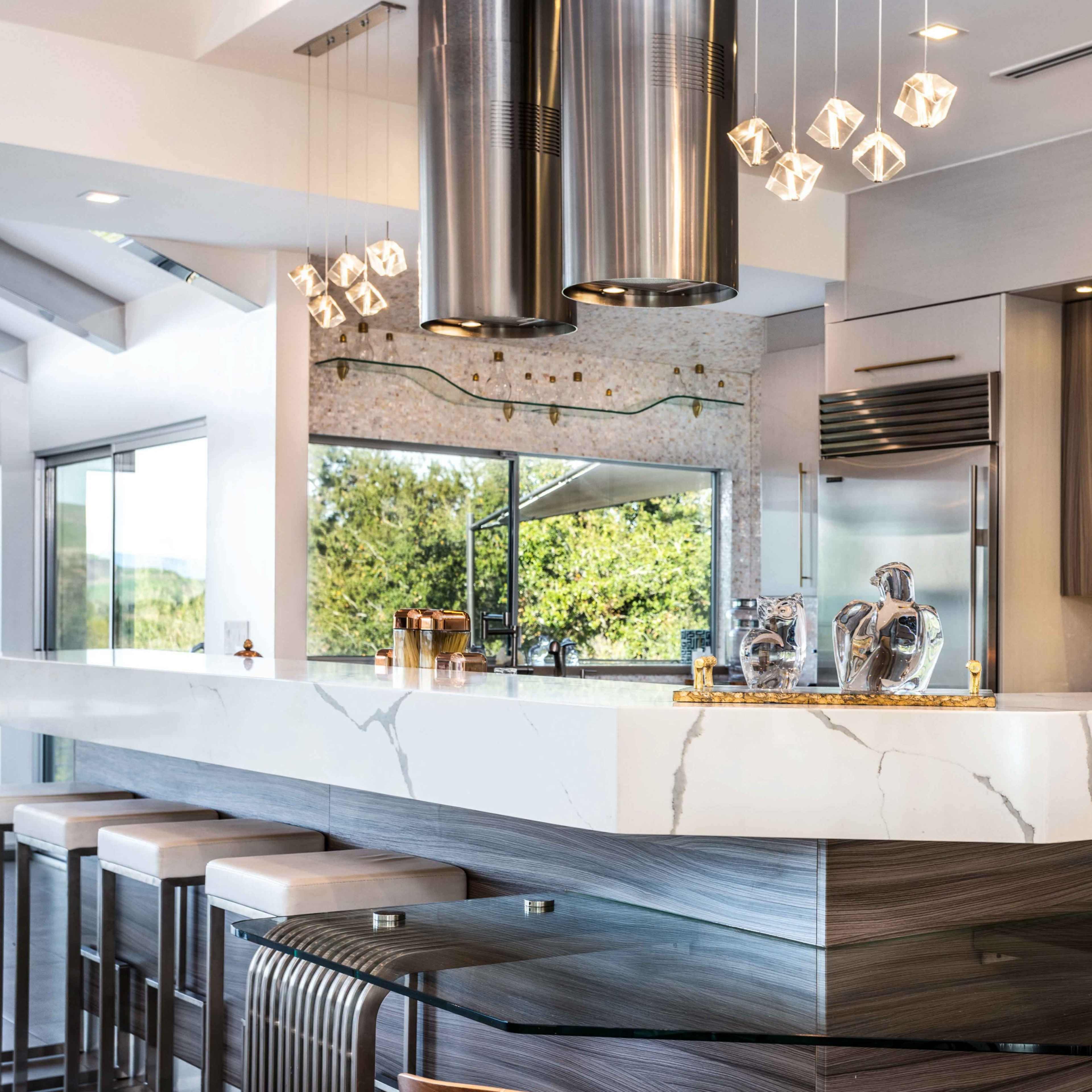 A modern kitchen features a large white marble countertop with metallic bar stools and stainless steel appliances, surrounded by ample natural light from large windows.