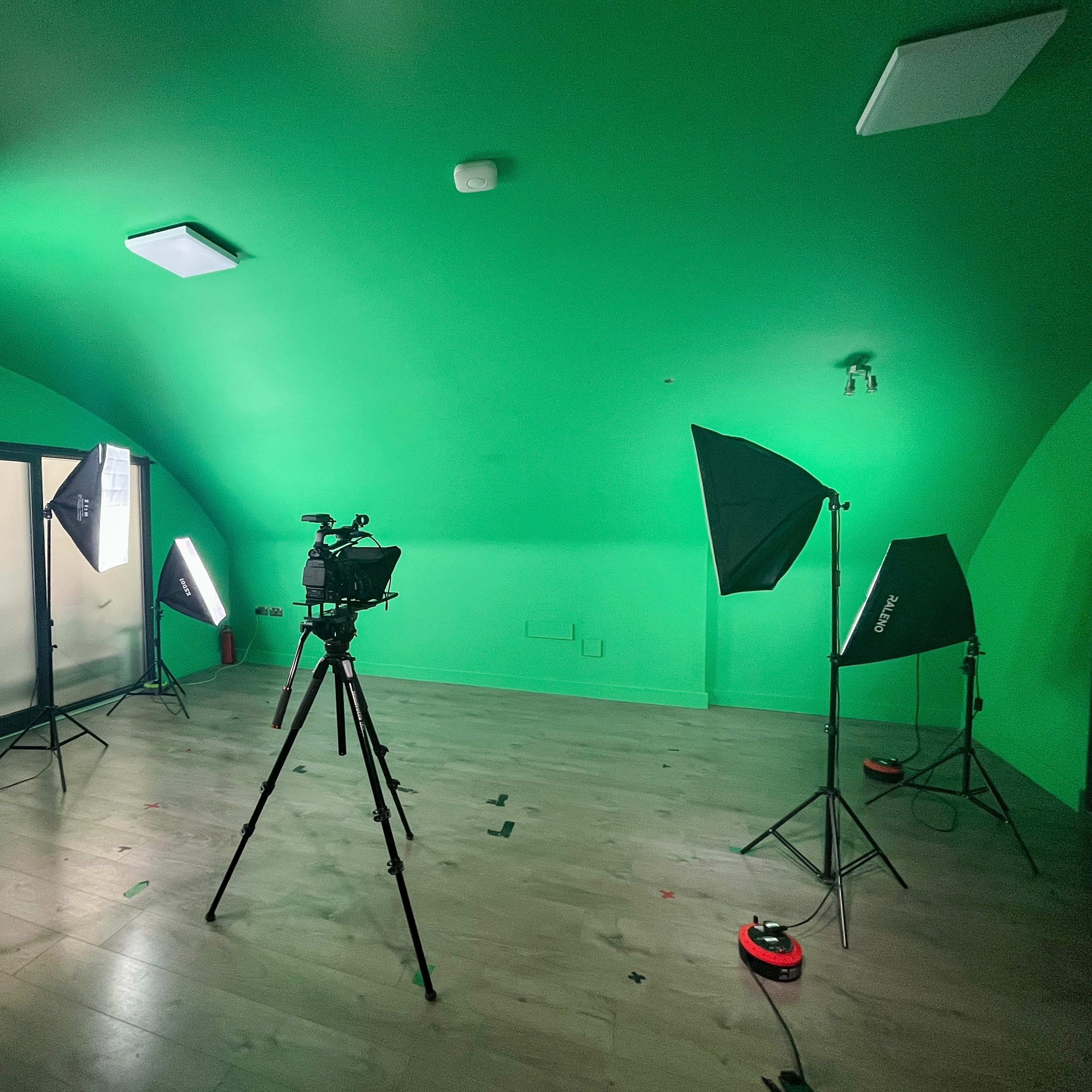 A green studio room is set up with lighting equipment and a camera on a tripod.