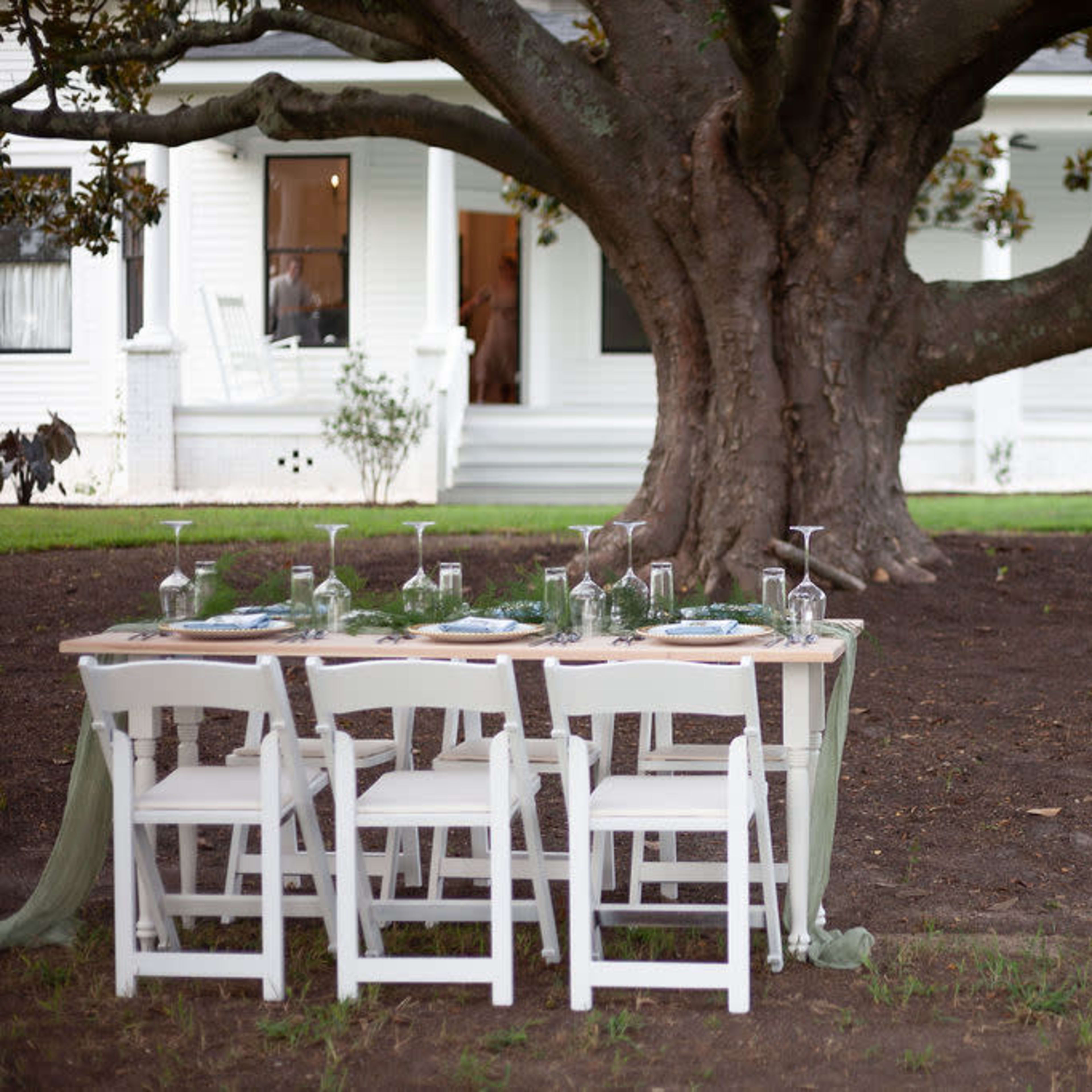 A dining table with white chairs is set under a large tree, near a white house in the background.