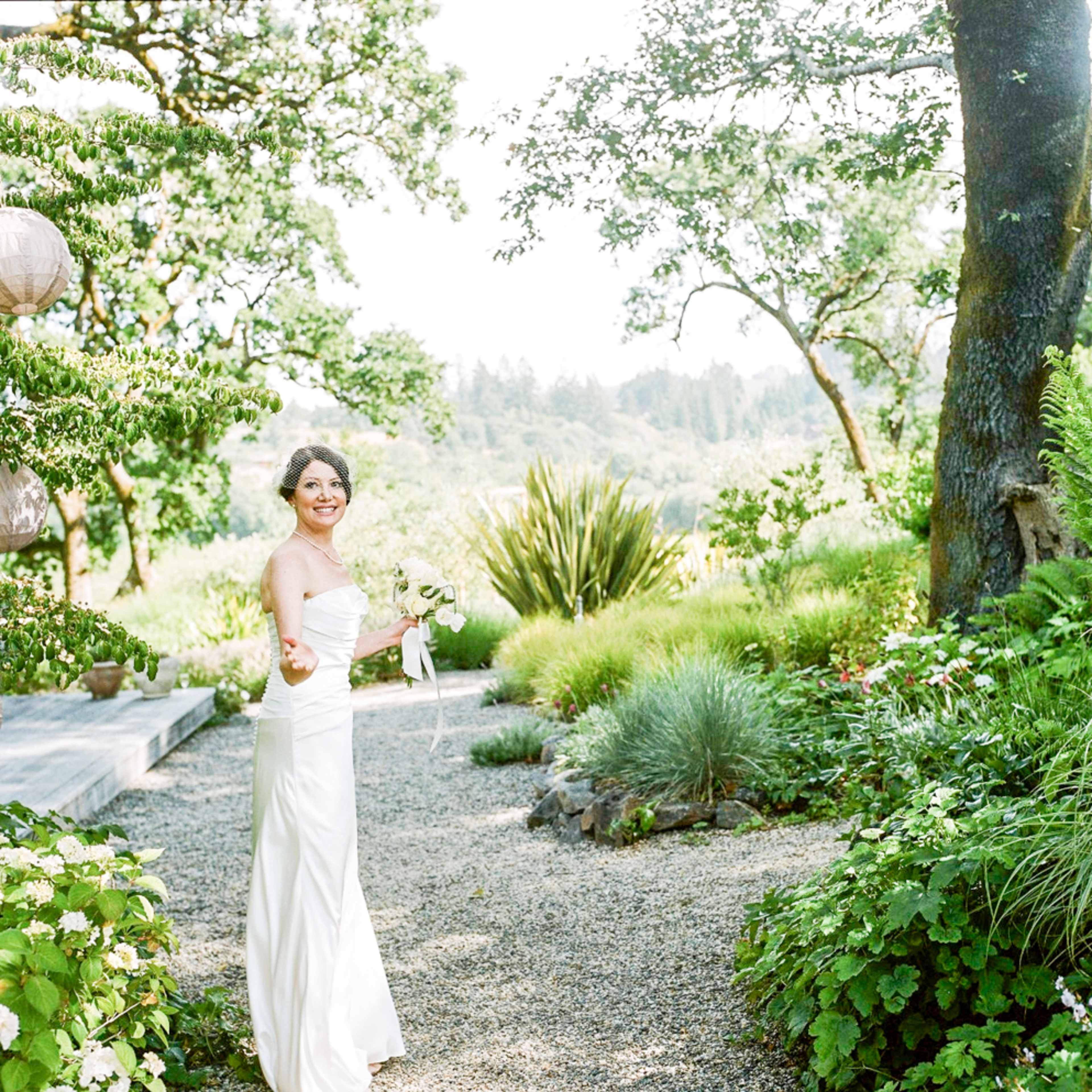 A woman in a white wedding dress stands in a garden filled with greenery, holding a bouquet and smiling.
