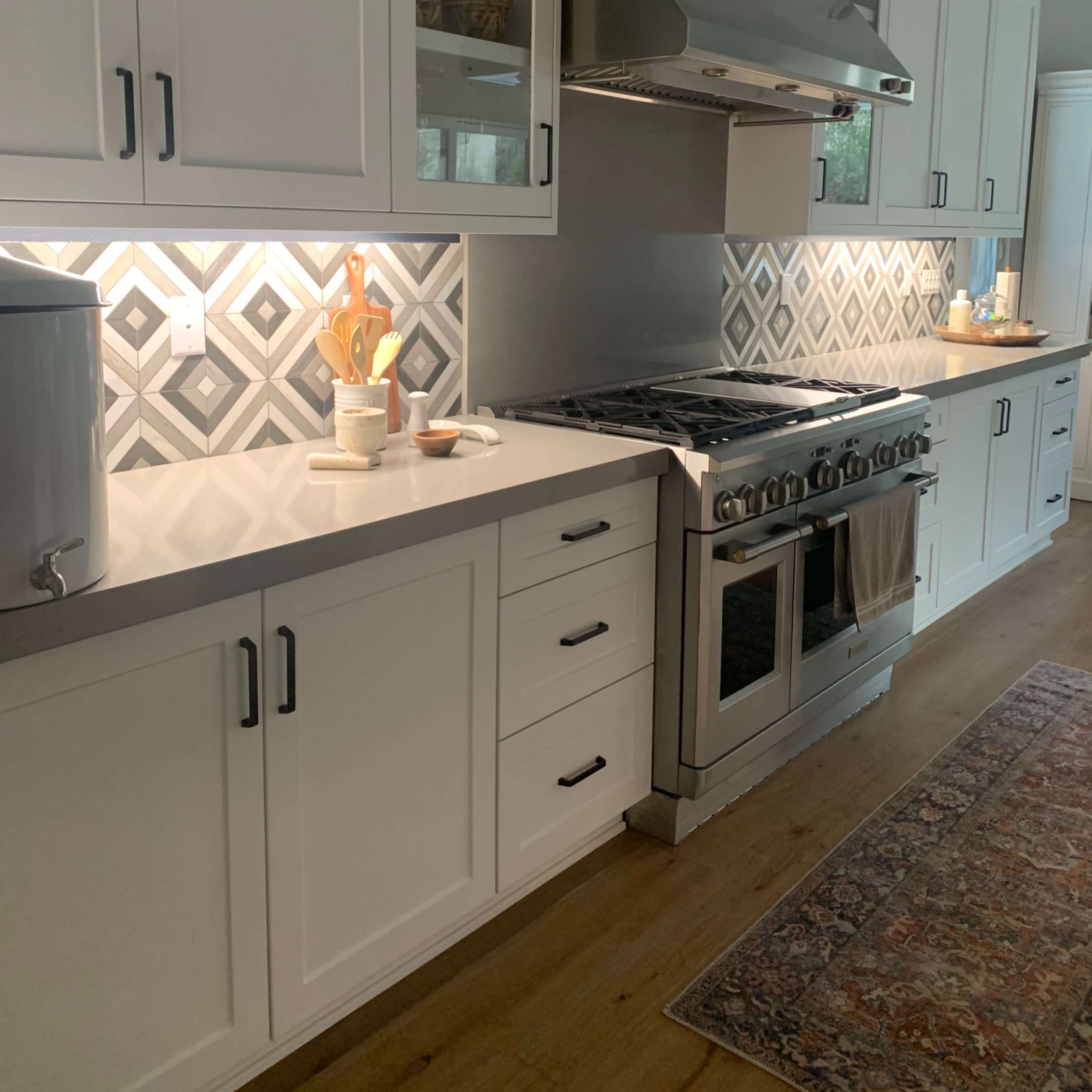 A modern kitchen featuring white cabinetry, a stainless steel range hood, and a patterned backsplash with warm lighting under the cabinets.