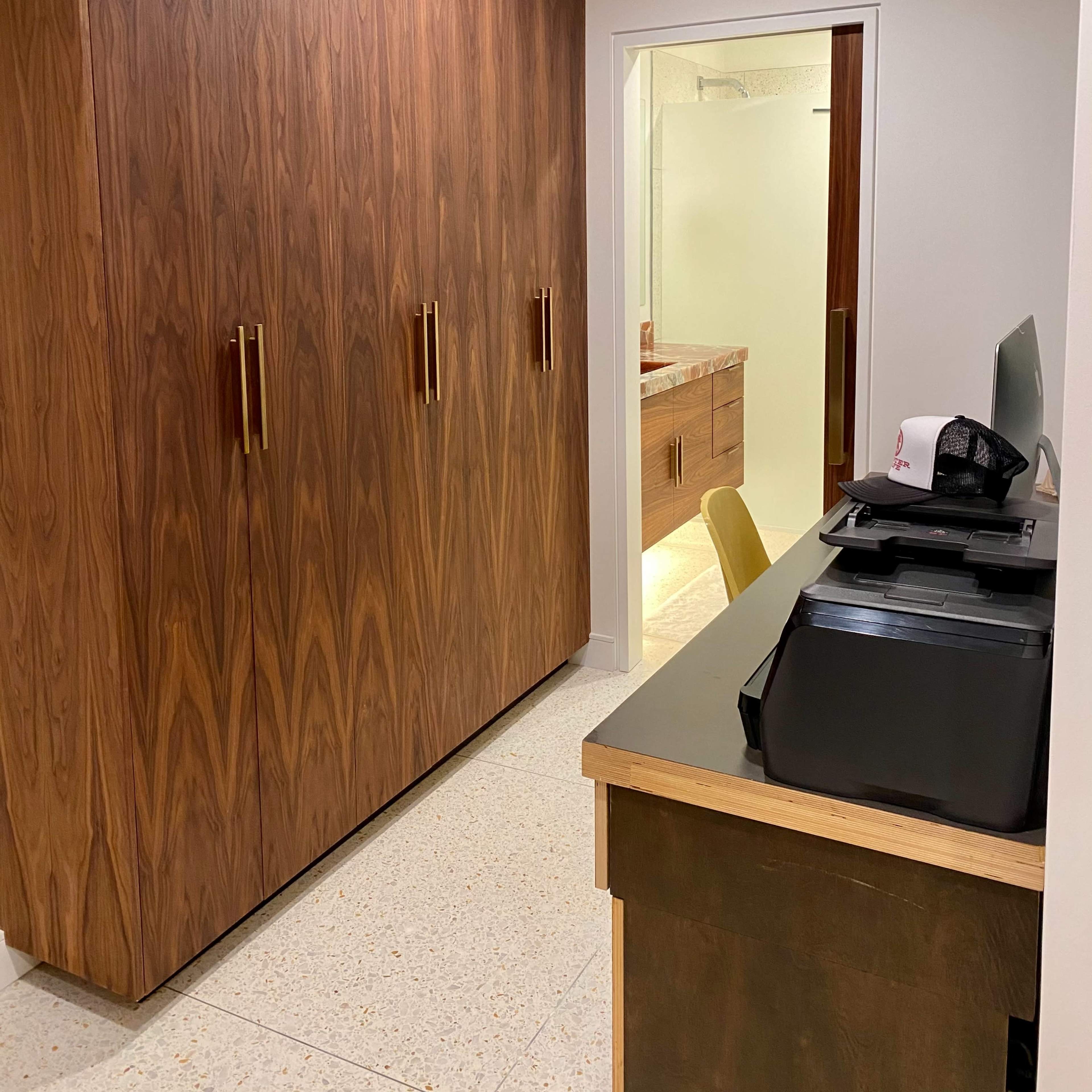 A room with wooden cabinetry on one side and a desk with a printer and a chair on the other, leading to a bathroom in the background.