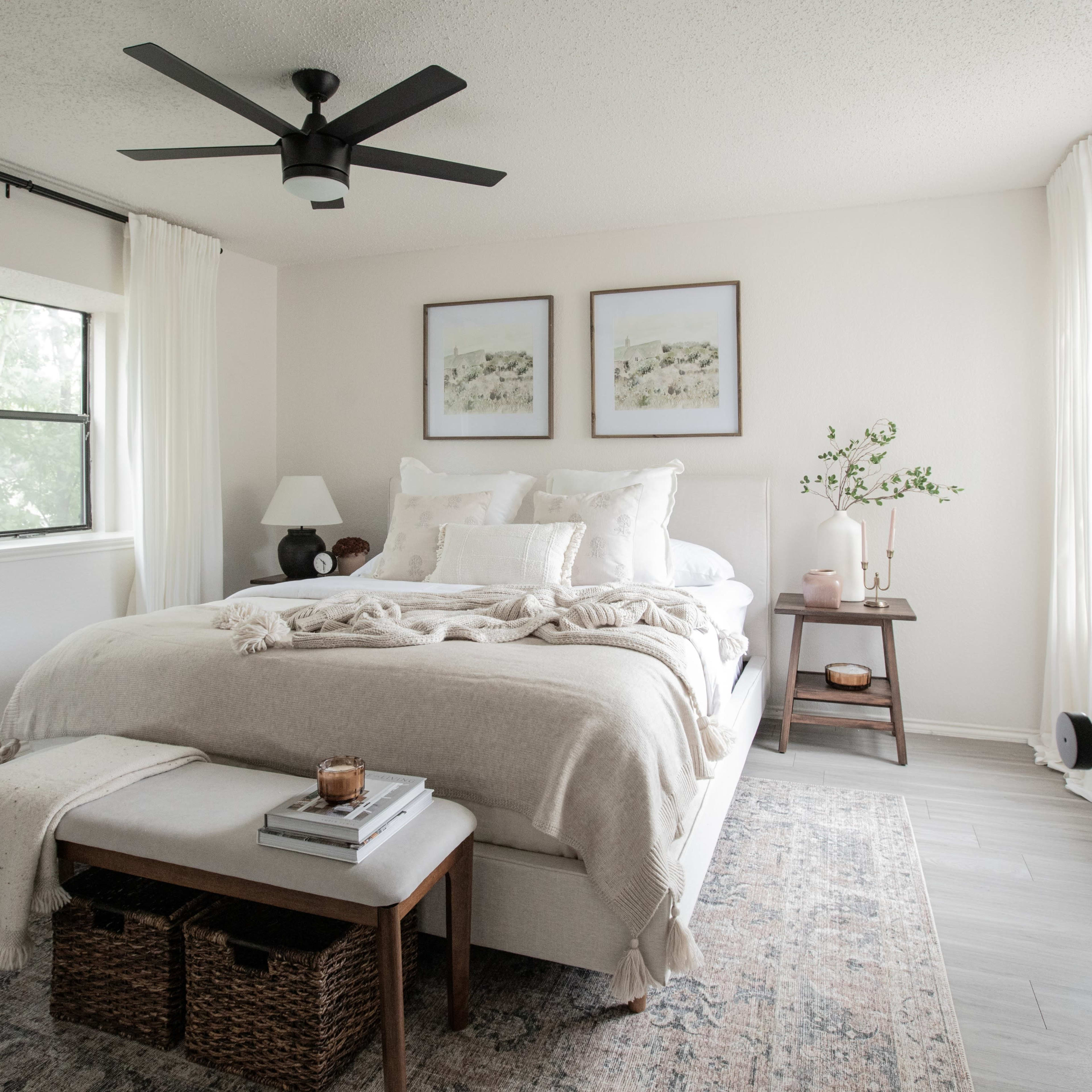 A well-lit bedroom features a large bed with white bedding, a bedside table with a lamp, and framed artwork on the walls.