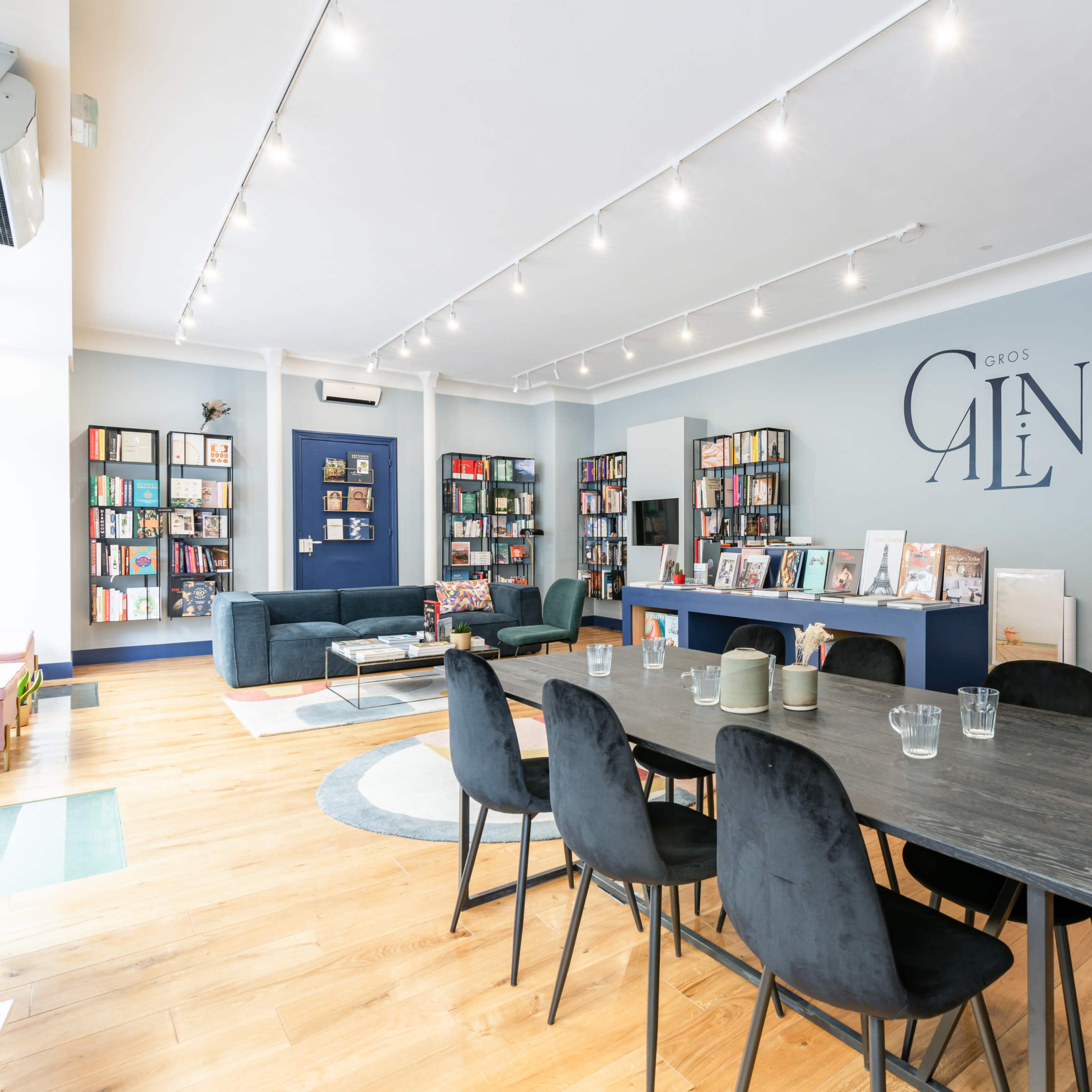 A modern, well-lit community space with bookshelves lining the walls, a large wooden dining table surrounded by black chairs, and comfortable seating areas.
