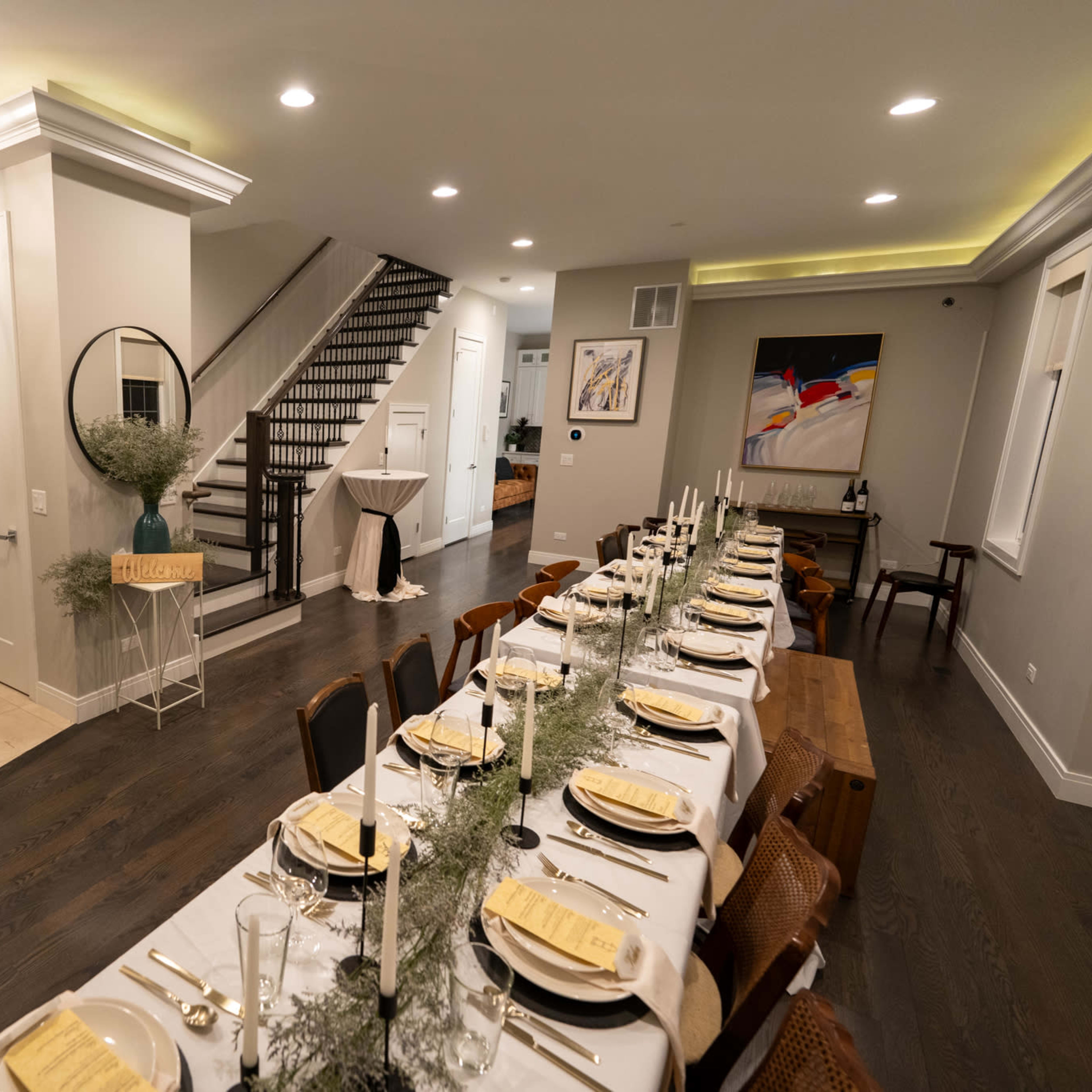A long dining table is set for a meal in a modern, well-lit room that features a staircase leading to another level.