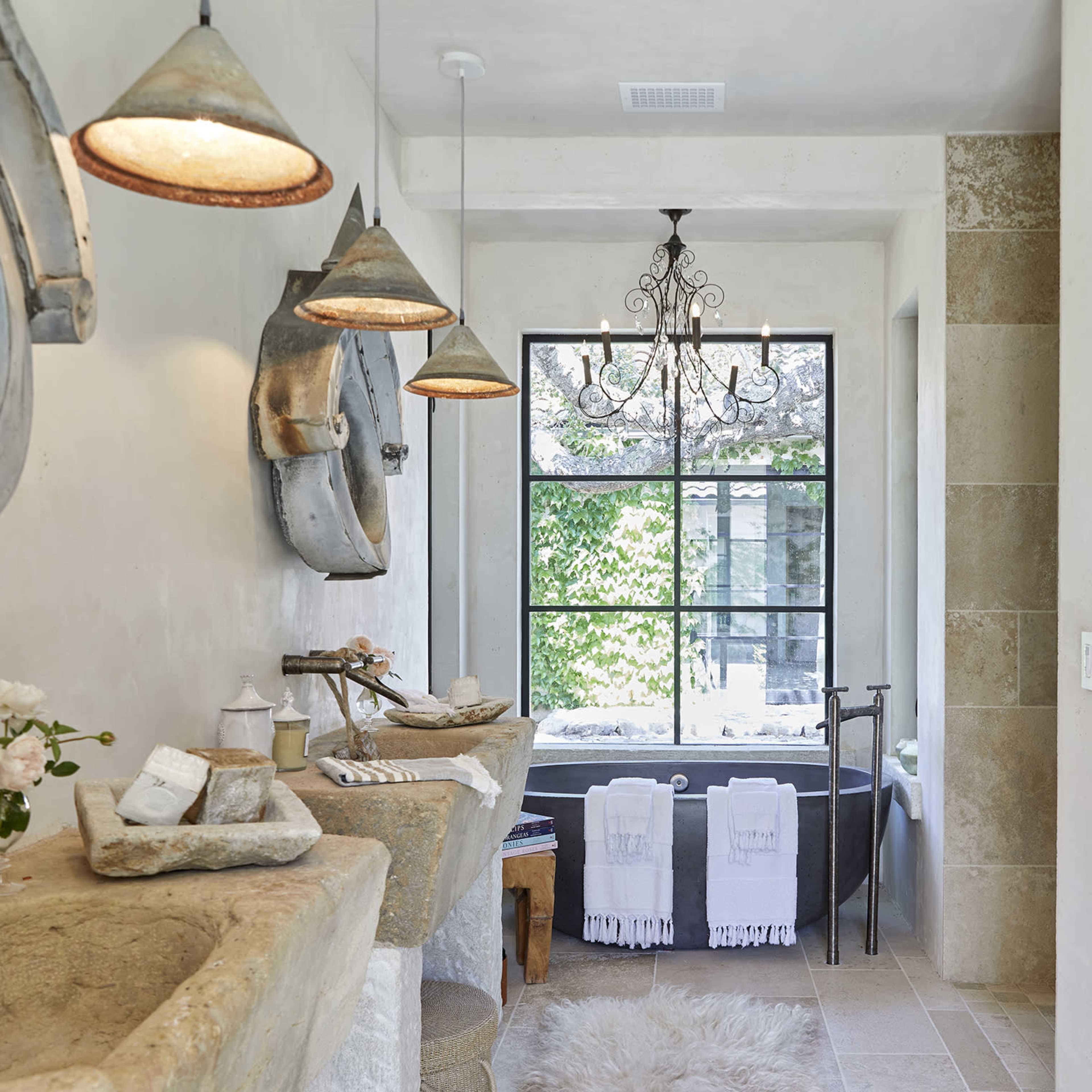 The bathroom features stone sinks, vintage mirrors, a chandelier, and a large window overlooking an outdoor area.