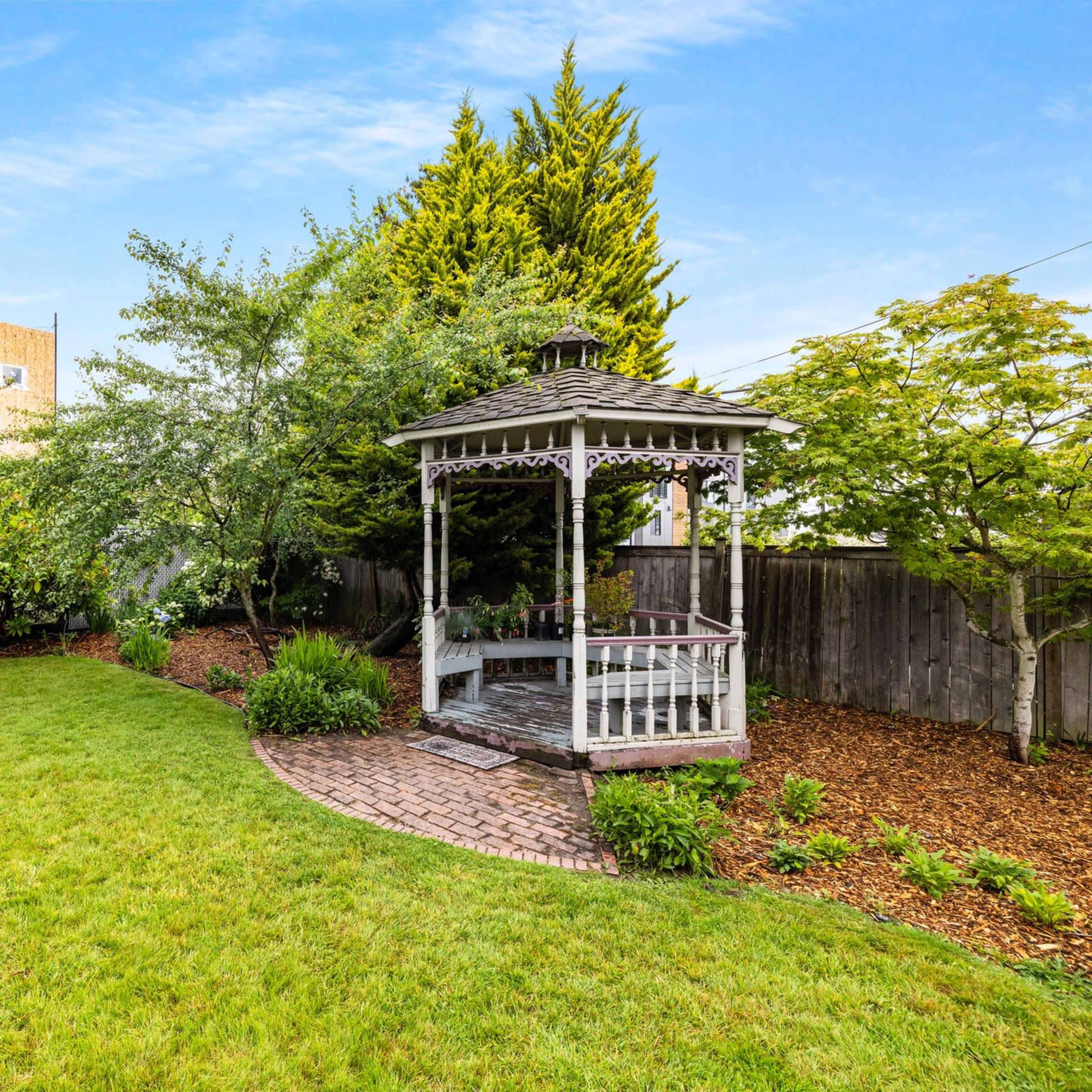 A landscaped backyard featuring a gazebo, lush green grass, and various trees and flowers.