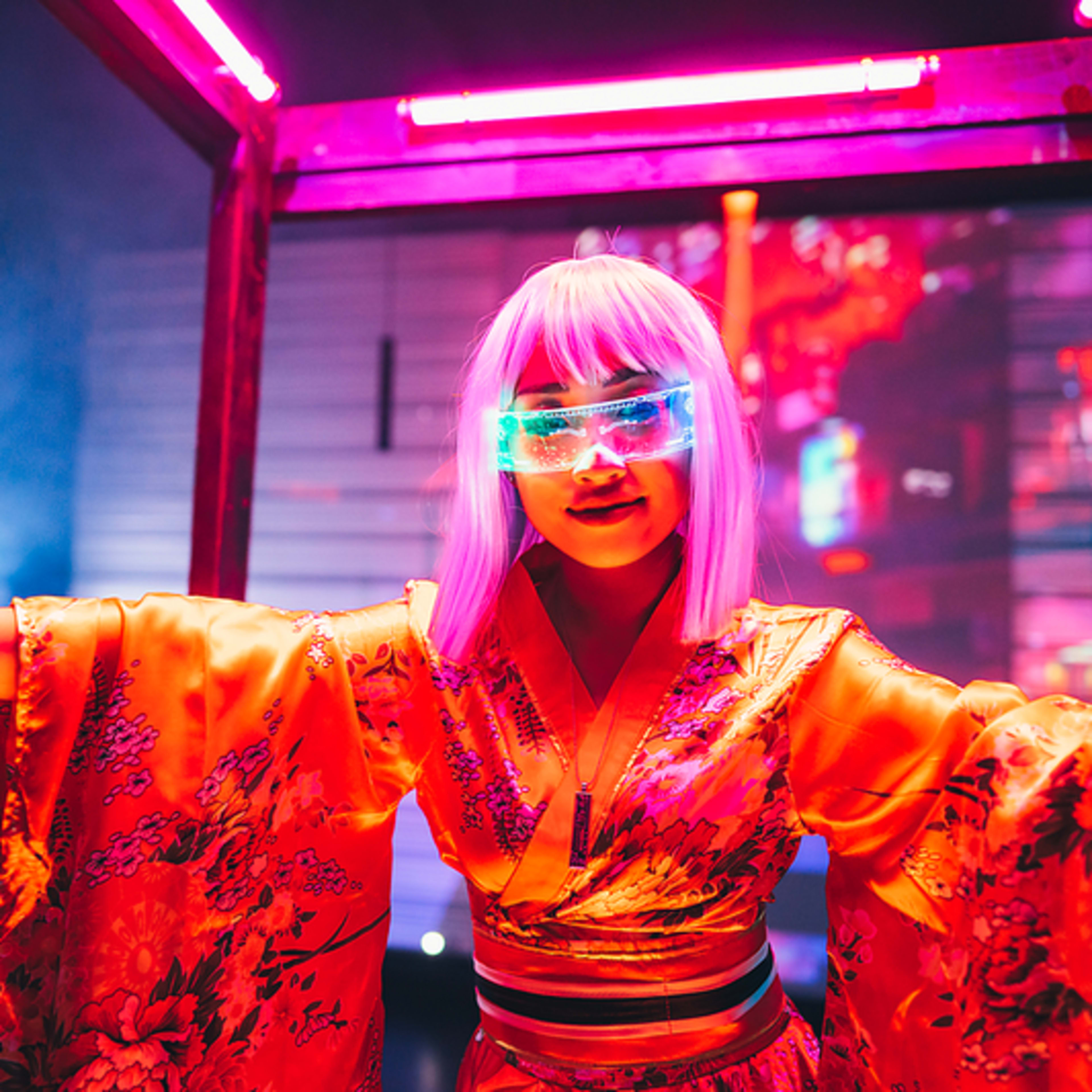 A person wearing a colorful kimono and futuristic glasses poses inside a neon-lit frame with a projected backdrop.