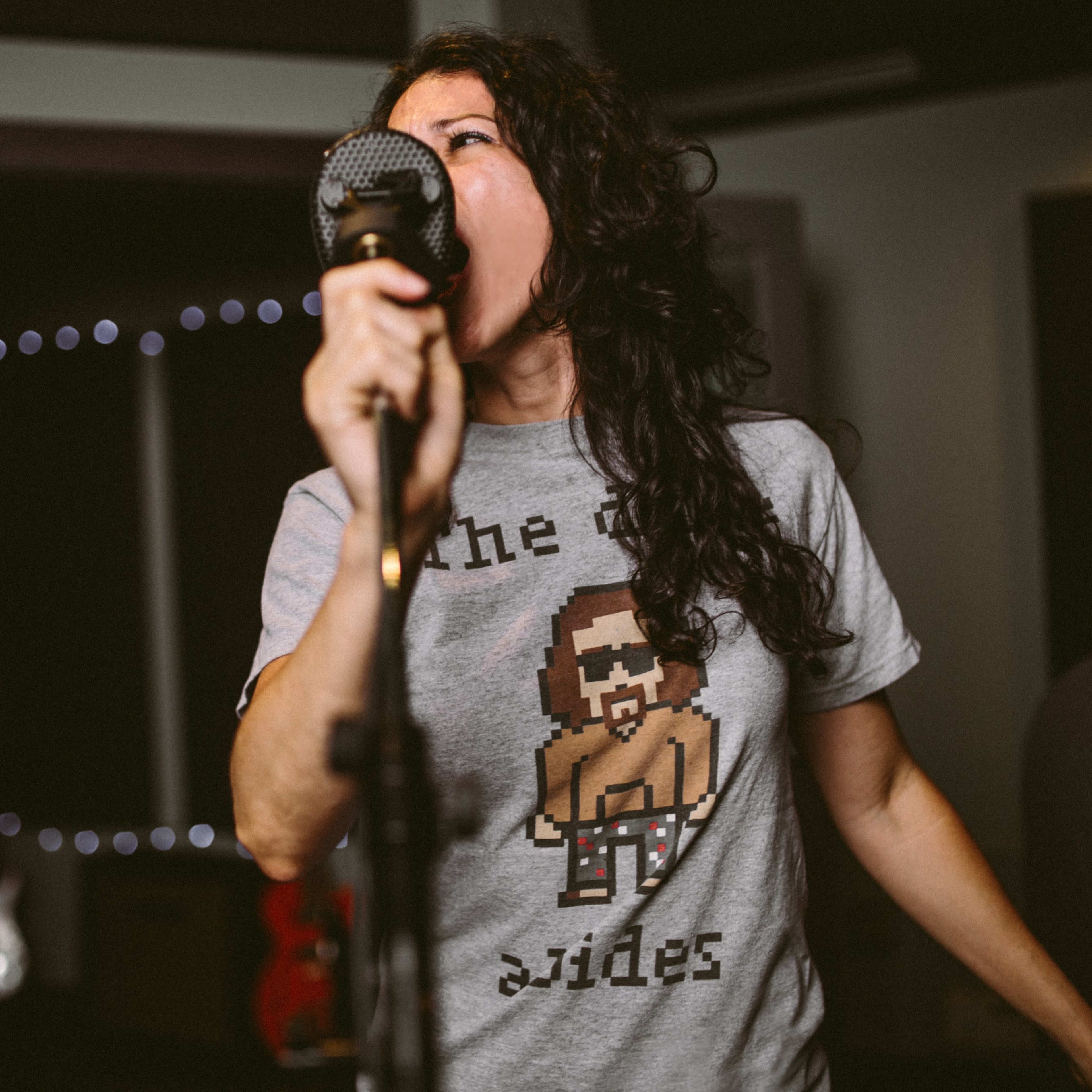 A woman sings into a microphone while wearing a graphic t-shirt featuring a pixelated character.