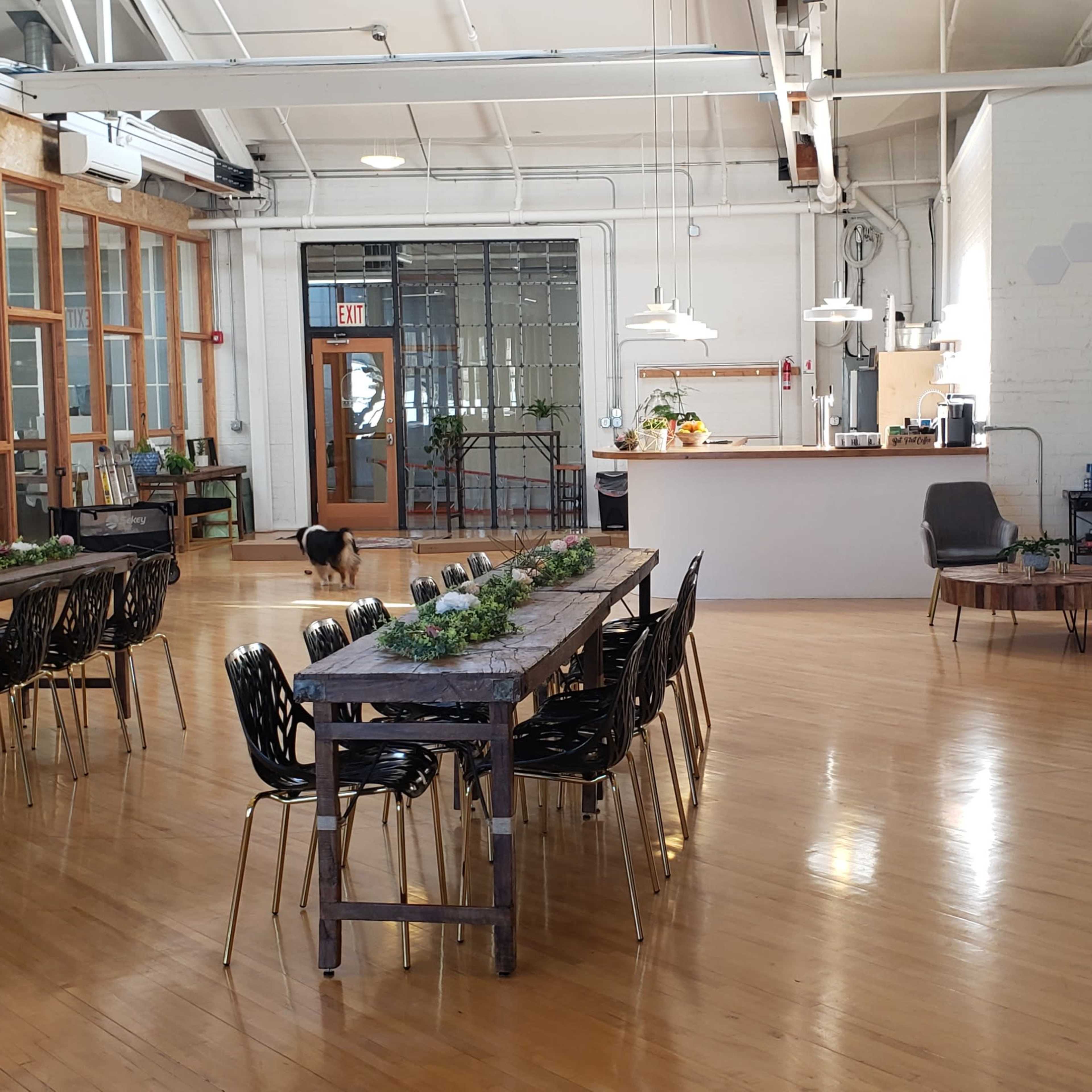 A spacious, modern event venue with wooden floors, long tables set for dining, and a large serving area in the background.