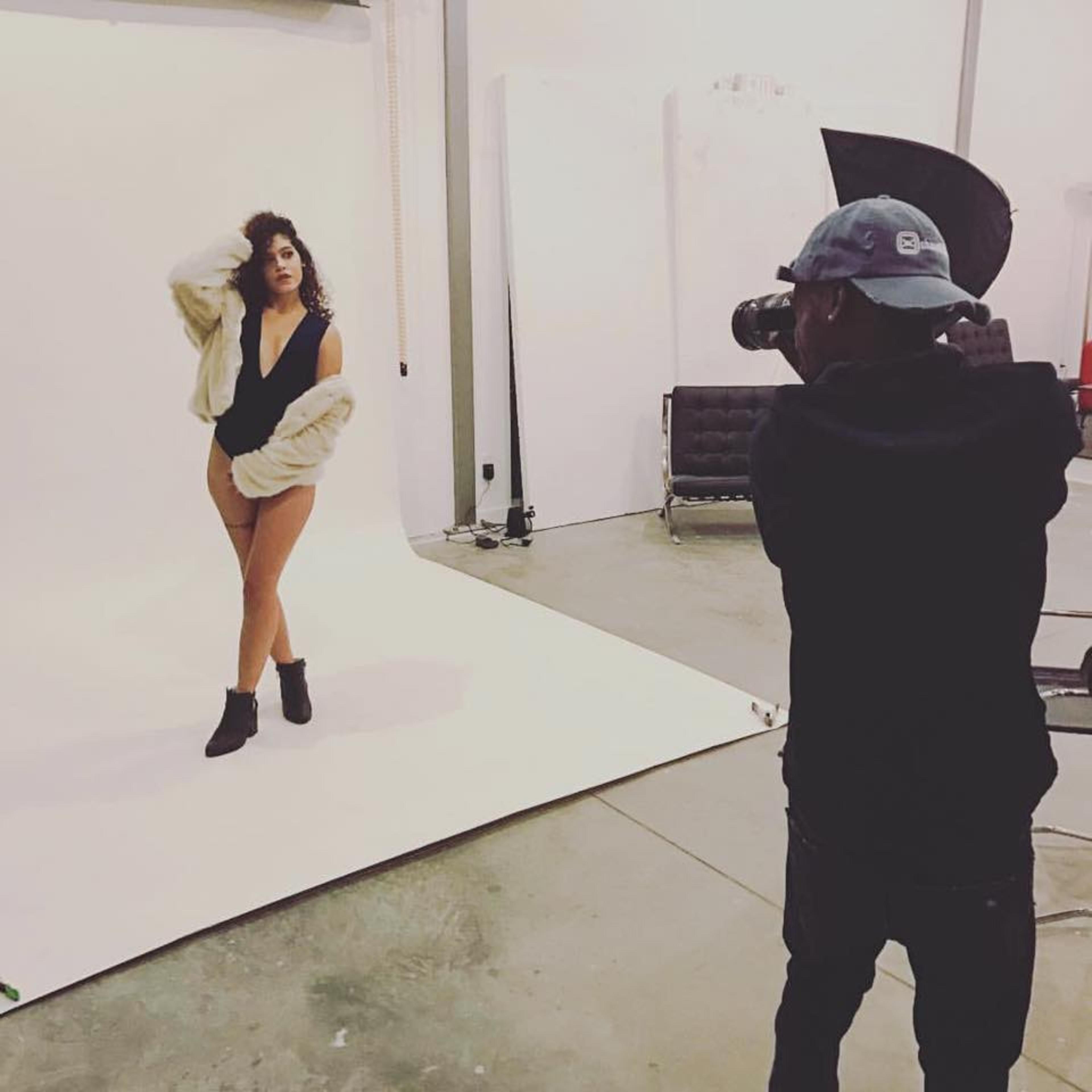 A photographer captures an image of a model posing in a studio with a white backdrop.